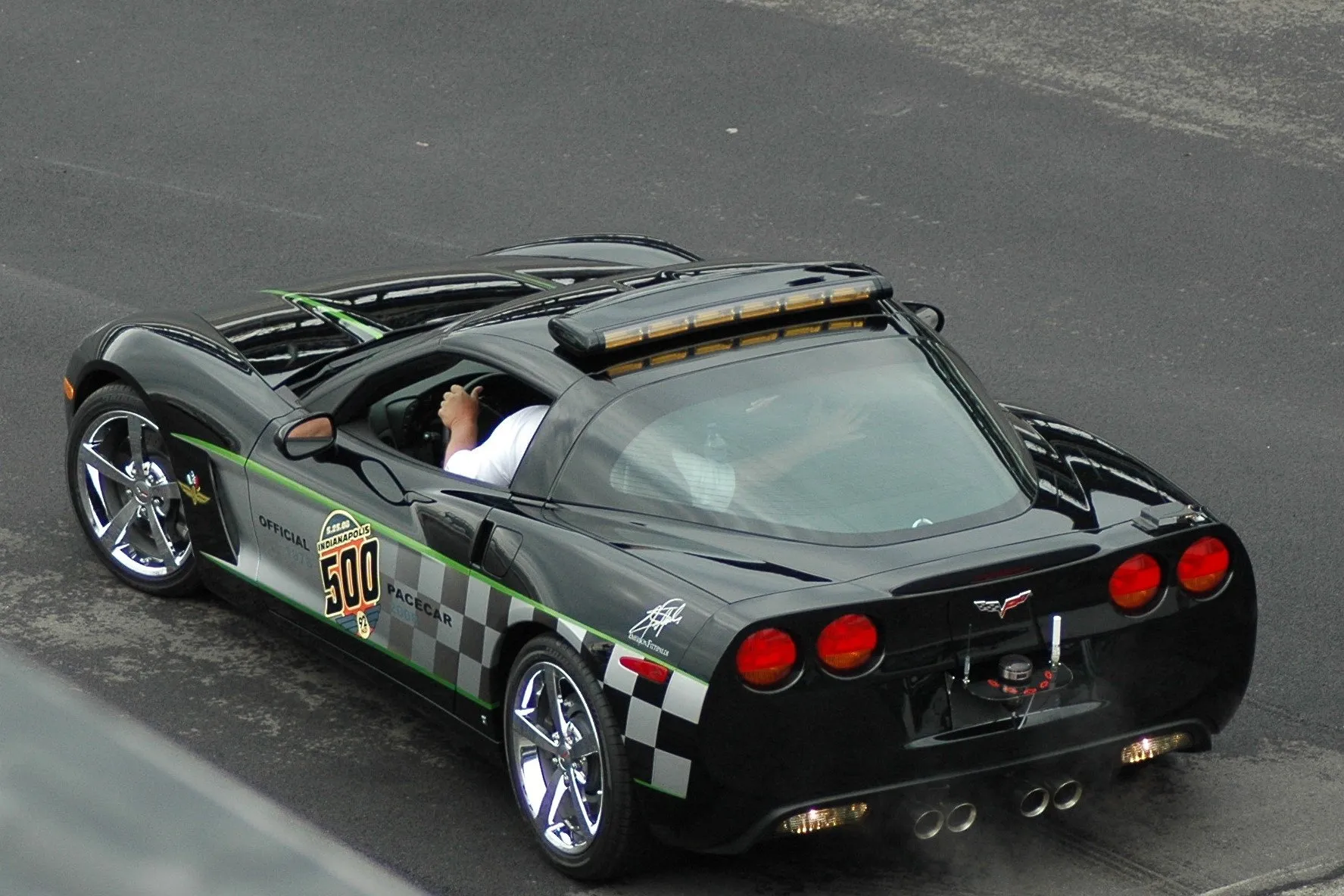 A black Corvette coupe seen from above and behind, with a light bar on the roof, a driver's arm at the open window, a 500 logo on the door and checkered graphics on the rear fender.