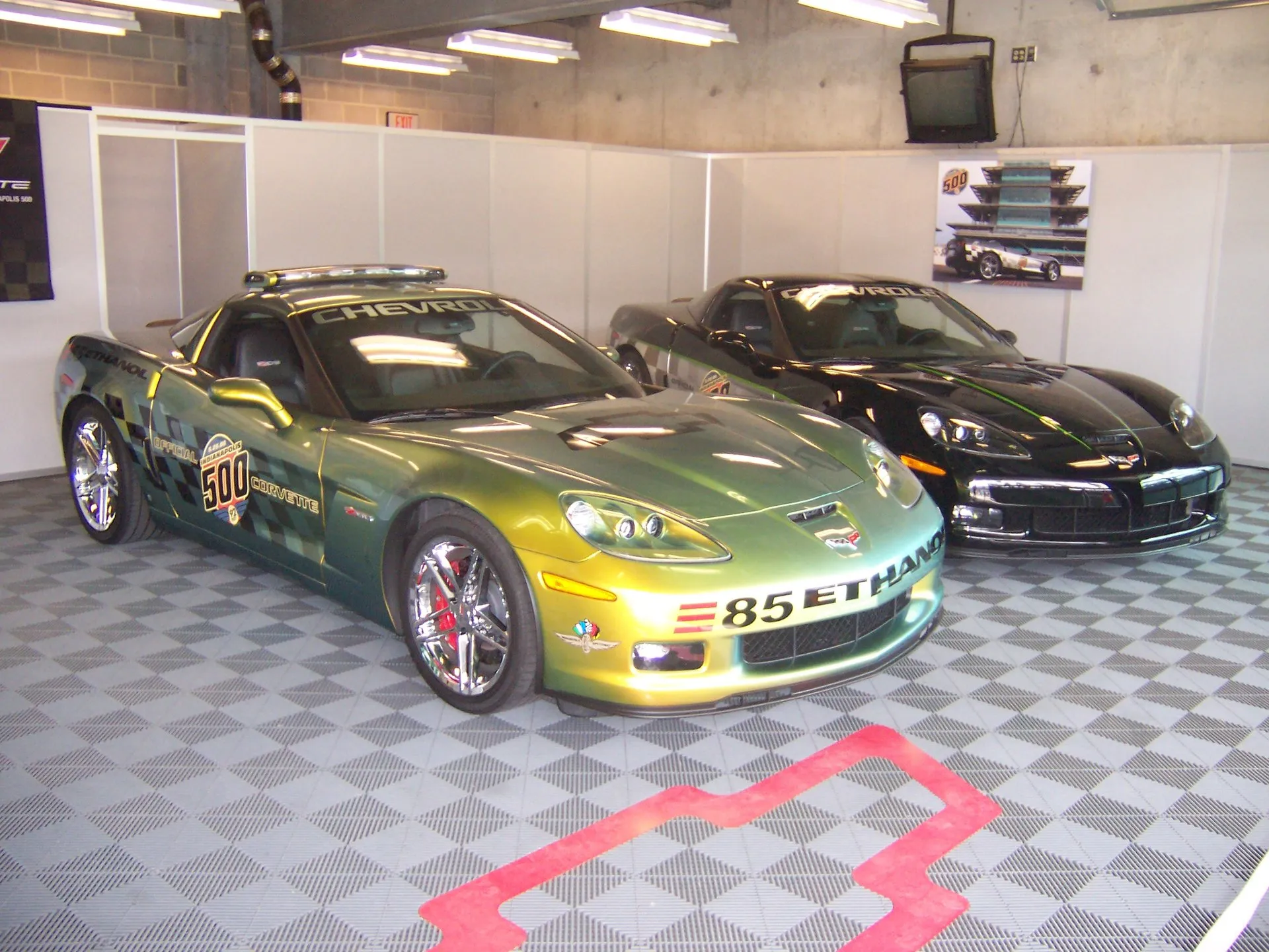 Two Corvettes in a garage with a checkered floor: a green and gold Z06 lettered E85 Ethanol in front, and a black coupe with a thin green stripe behind it.