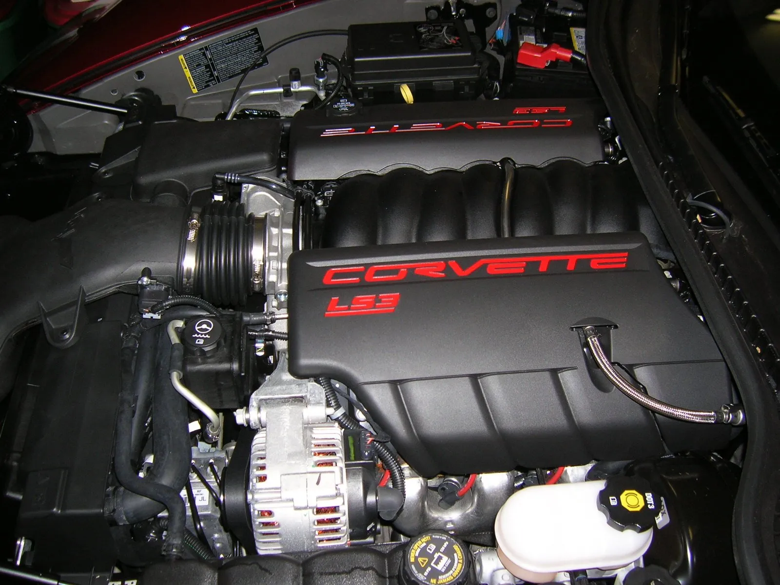 The engine bay of a C6 Corvette with the hood open, showing black engine covers lettered Corvette LS3 in red and the air intake in front of them.