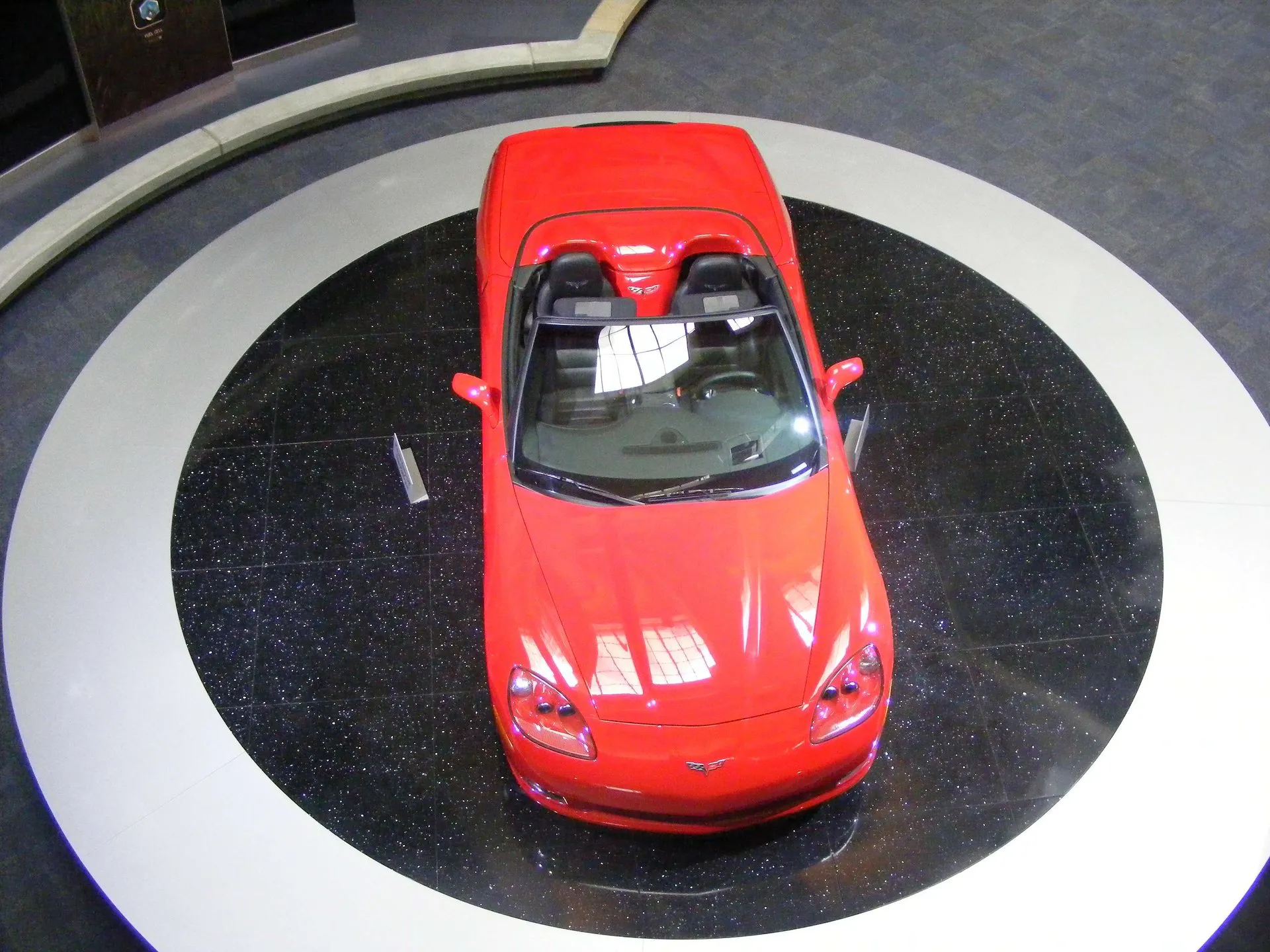 Red C6 Corvette convertible with its top down, seen from above on a round black and white display platform inside a building.