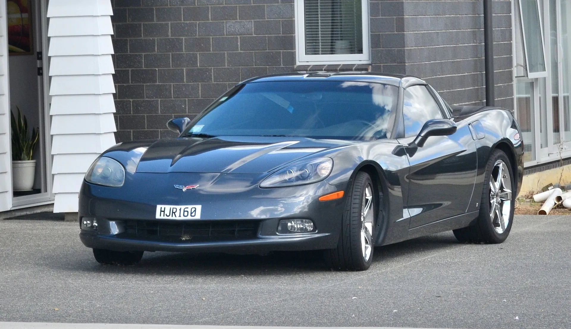Dark gray C6 Corvette coupe with chrome five-spoke wheels and a New Zealand number plate, parked on a driveway in front of a brick house, seen from the front left.