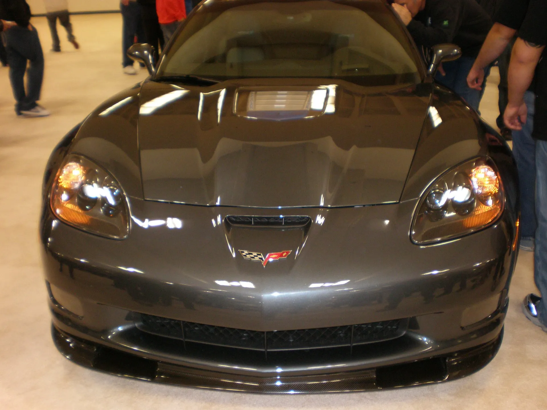 Dark gray C6 Corvette ZR1 on an auto show floor, seen head-on, with a clear window in the raised center of its hood, a carbon fiber front splitter and visitors standing beside it.