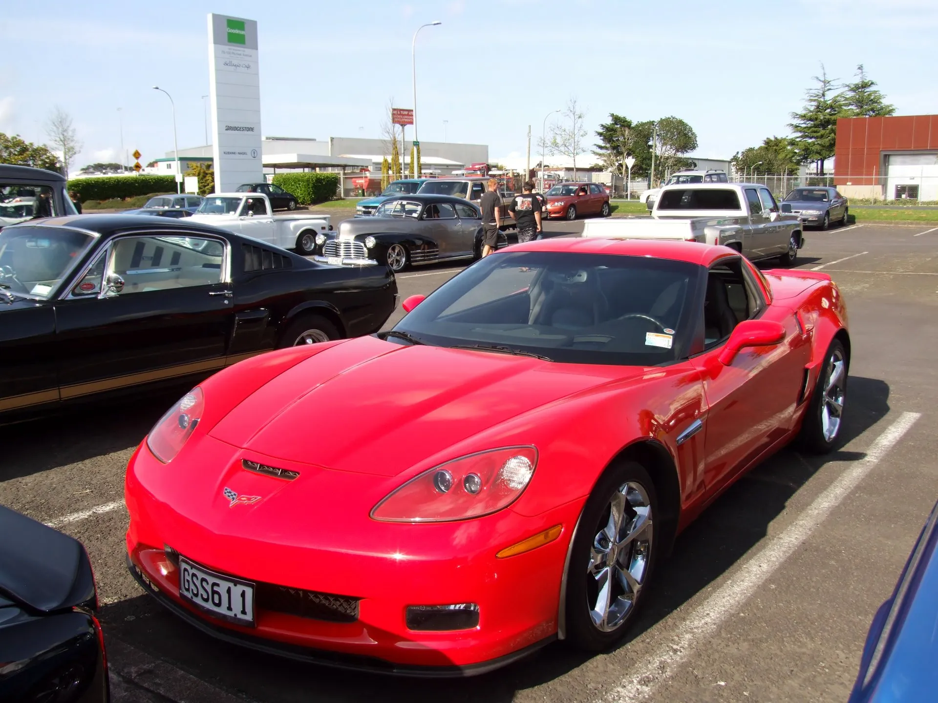Red C6 Corvette Grand Sport coupe with chrome wheels and a New Zealand number plate, parked at a car show beside a black classic fastback, seen from the front right.
