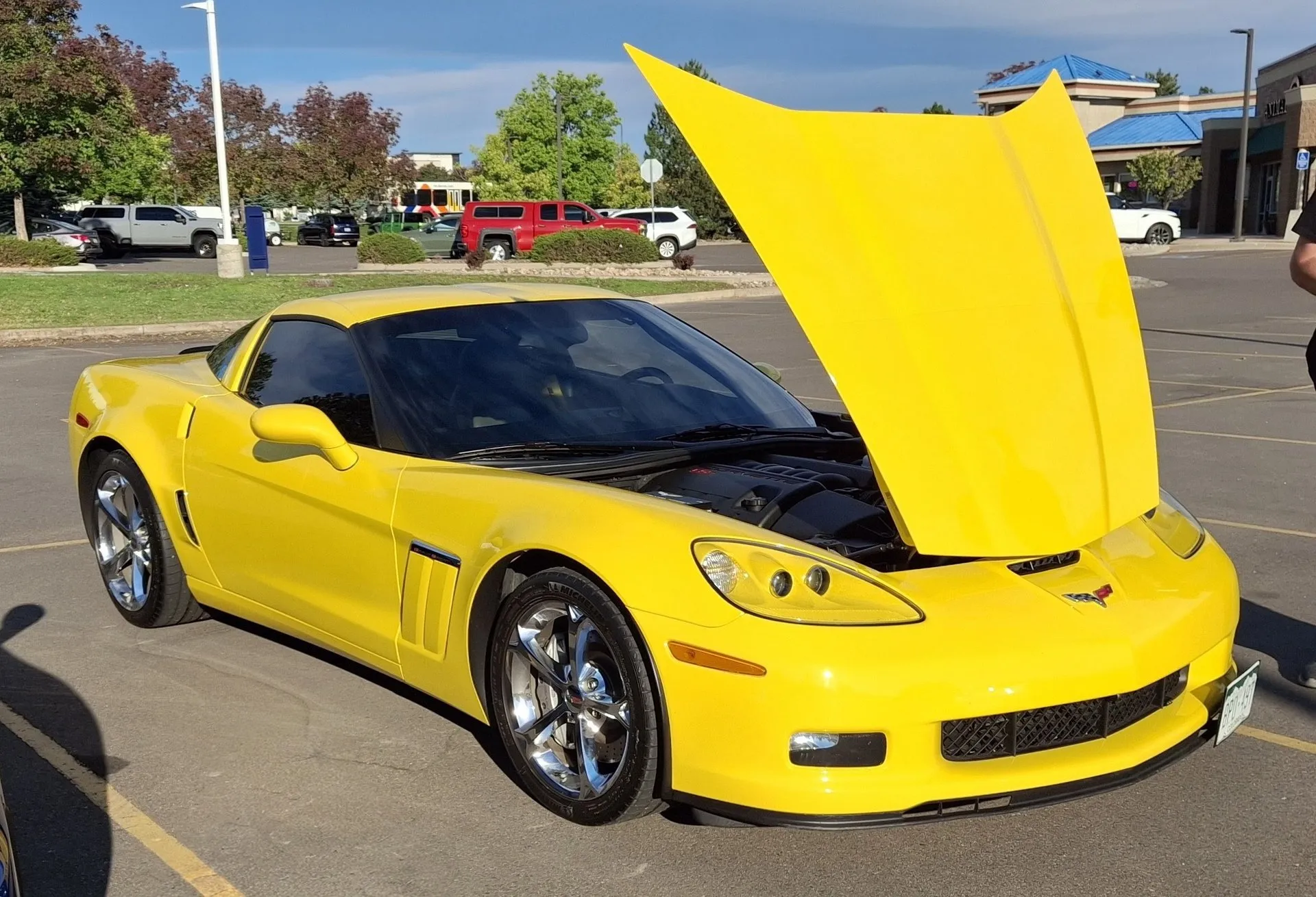 Yellow C6 Corvette Grand Sport coupe with its hood raised and chrome wheels, parked in a parking lot, seen from the front left, showing the wide front fender and its vent badge.