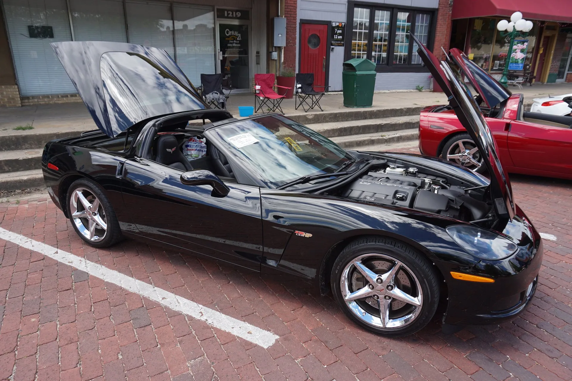 Black C6 Corvette convertible with its top stowed, hood and the lid behind the seats raised, on five-spoke chrome wheels, parked on a brick street.