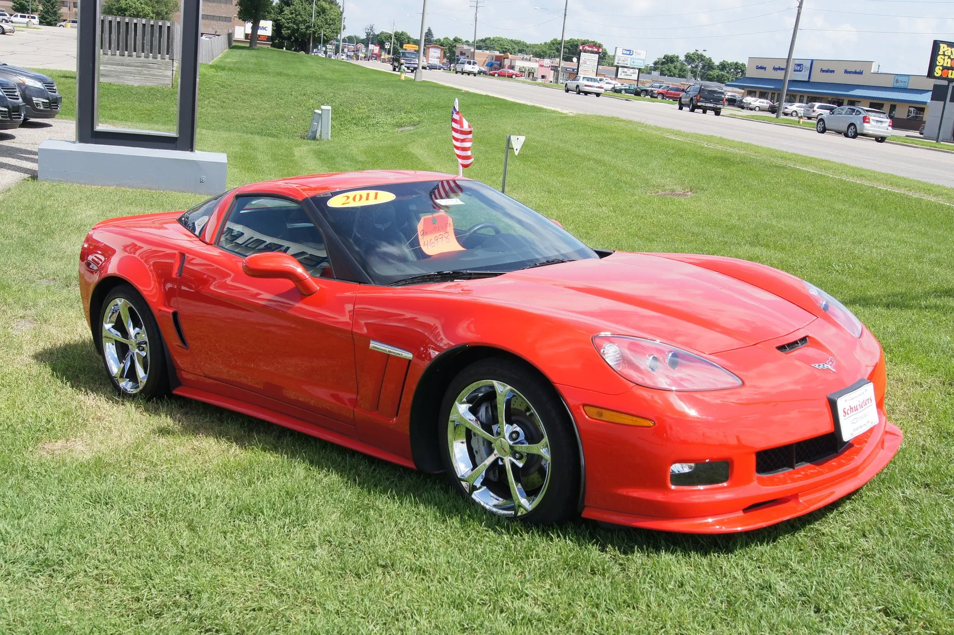 Red C6 Corvette Grand Sport coupe with chrome wheels on the grass of a car dealer's lot, seen from the front right, with a yellow 2011 sticker and a price tag on the windshield.