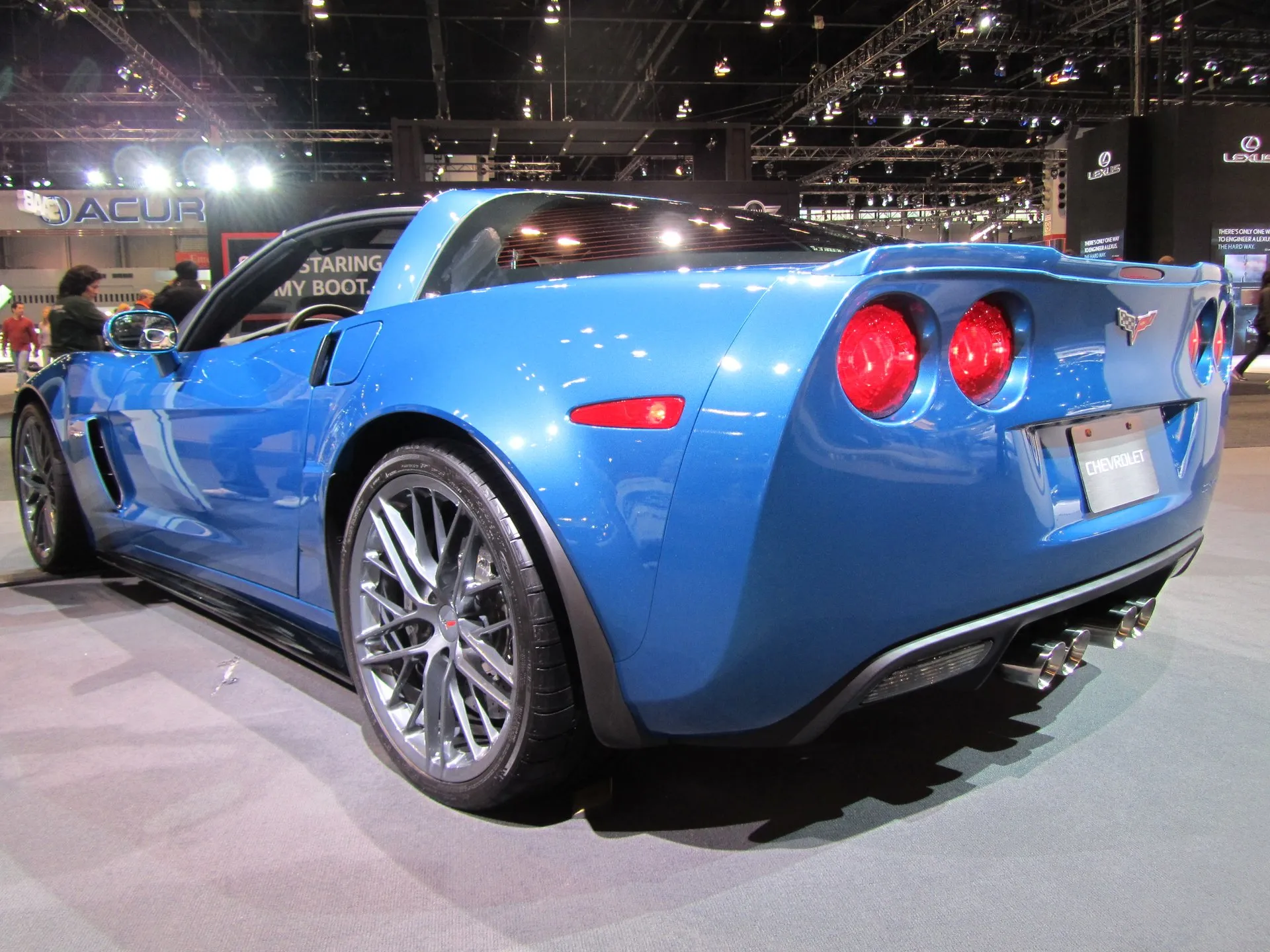 Bright blue C6 Corvette Z06 coupe on an auto show stand, seen from the rear left, with gray 20-spoke wheels, round red taillights and four exhaust tips.