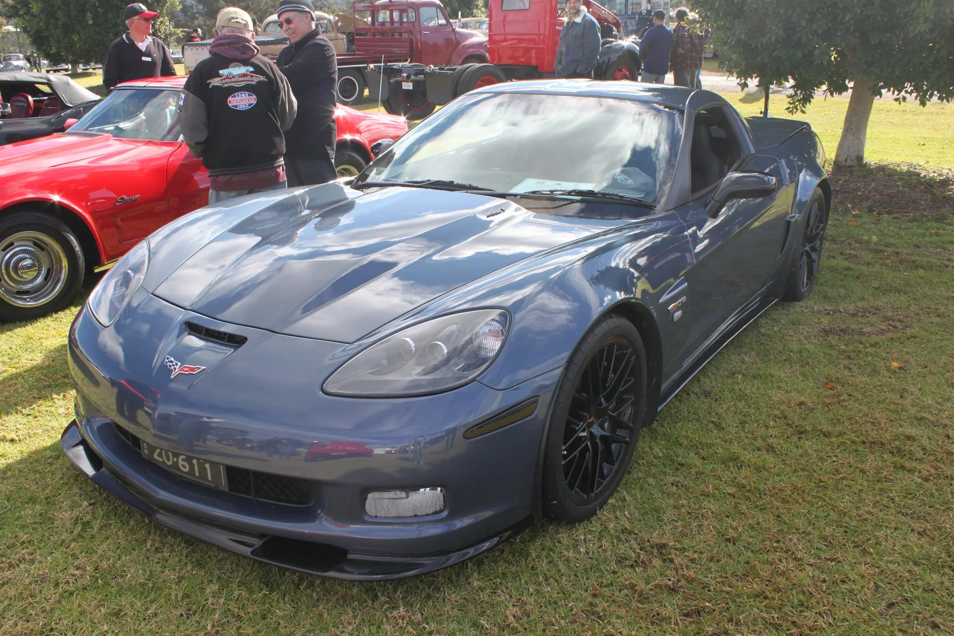 Blue-gray 2011 Chevrolet Corvette Z06 on grass at an outdoor car show in Australia, seen from the front left, with black multi-spoke wheels, a raised center section on the hood and a red C3 Corvette parked beside it.