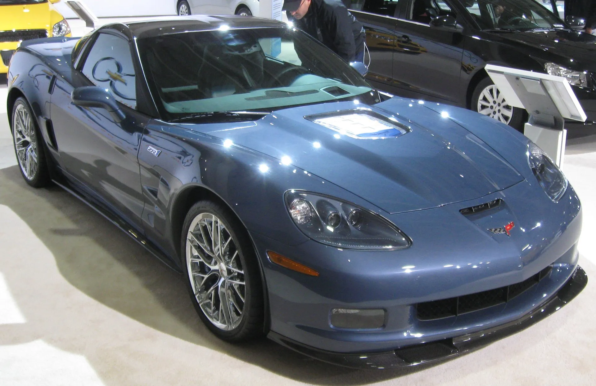 Blue-gray C6 Corvette ZR1 coupe with a clear window in its hood, a carbon fiber splitter and silver multi-spoke wheels, on display at an indoor auto show.