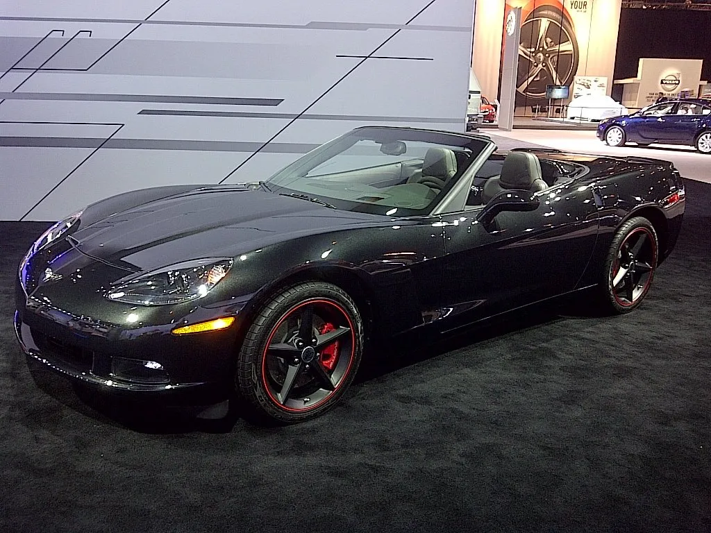 Dark gray metallic Corvette convertible with the top down on an auto show stand, with satin black hood stripes, black five-spoke wheels outlined in red and red brake calipers.