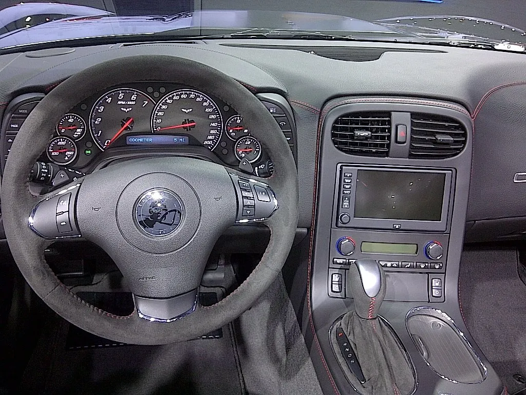 Driver's view of a Corvette Centennial Edition cabin: a suede-rimmed three-spoke steering wheel with a Louis Chevrolet medallion in the hub, the gauges, the navigation screen and an automatic shifter, with red stitching on the dash and console.