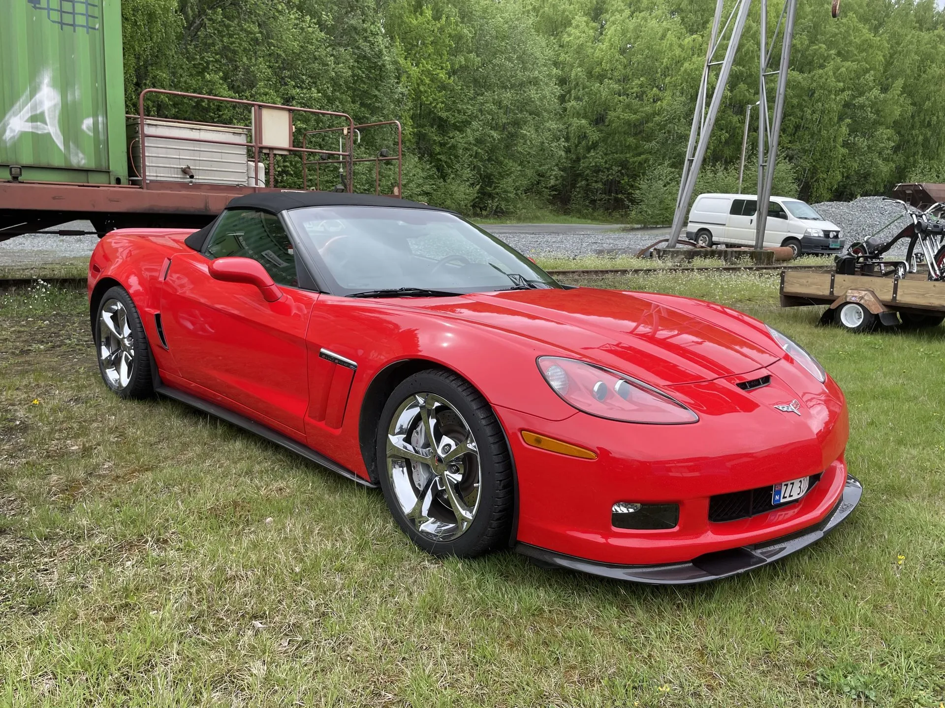Red Chevrolet Corvette Grand Sport convertible with a black soft top and chrome wheels, parked on grass beside an old rail wagon, seen from the front left.