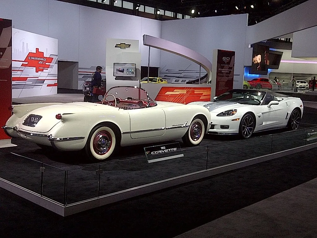 A white 1953 Corvette roadster with a red interior and whitewall tires parked beside a white C6 Corvette convertible with silver-blue stripes over its raised hood, on a Chevrolet auto show stand.