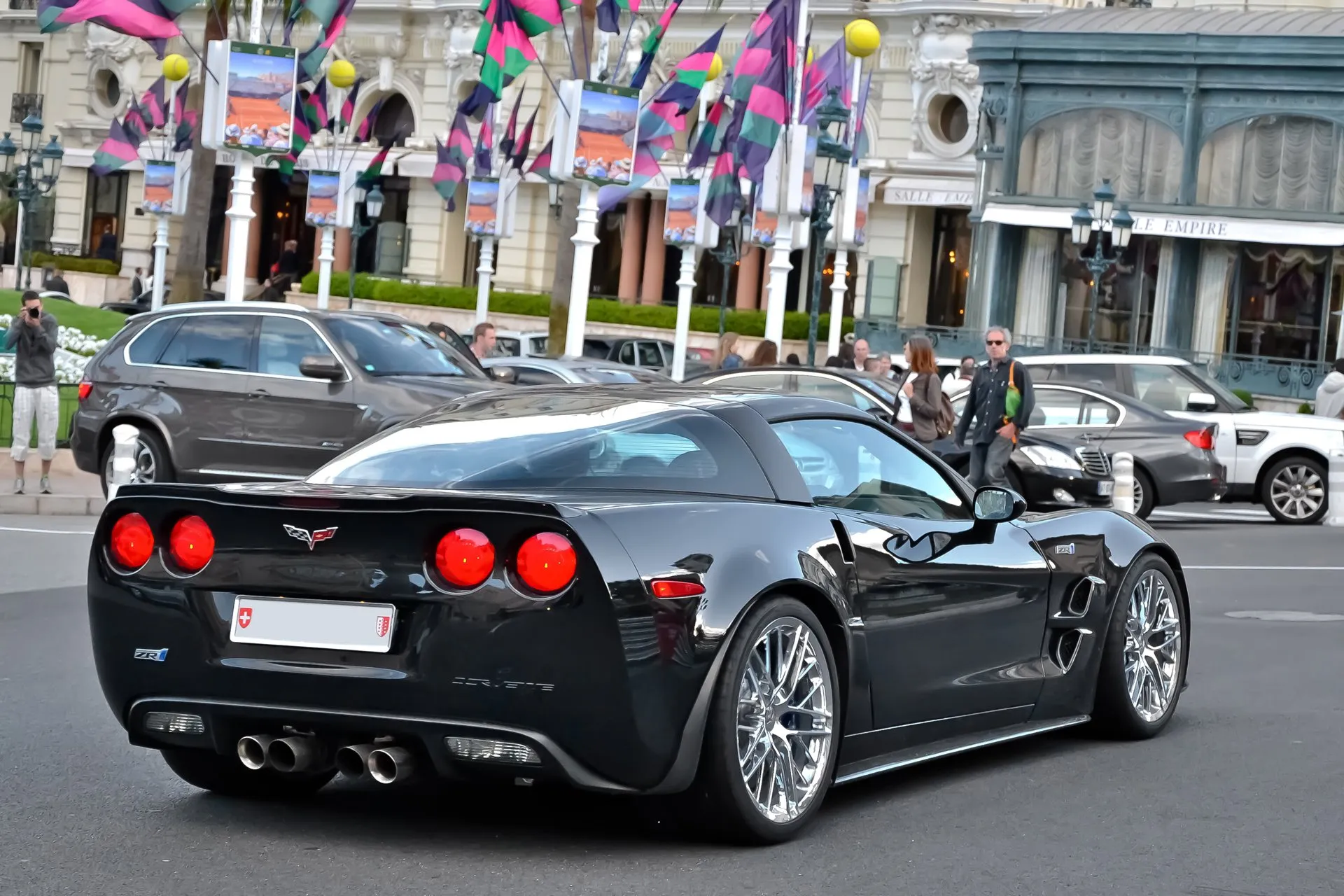 Black Corvette ZR1 seen from behind and to its right on a street in Monaco, showing a ZR1 badge on the rear panel, four round taillights, four exhaust tips, a rear spoiler and chrome multi-spoke wheels, with flags and buildings behind.