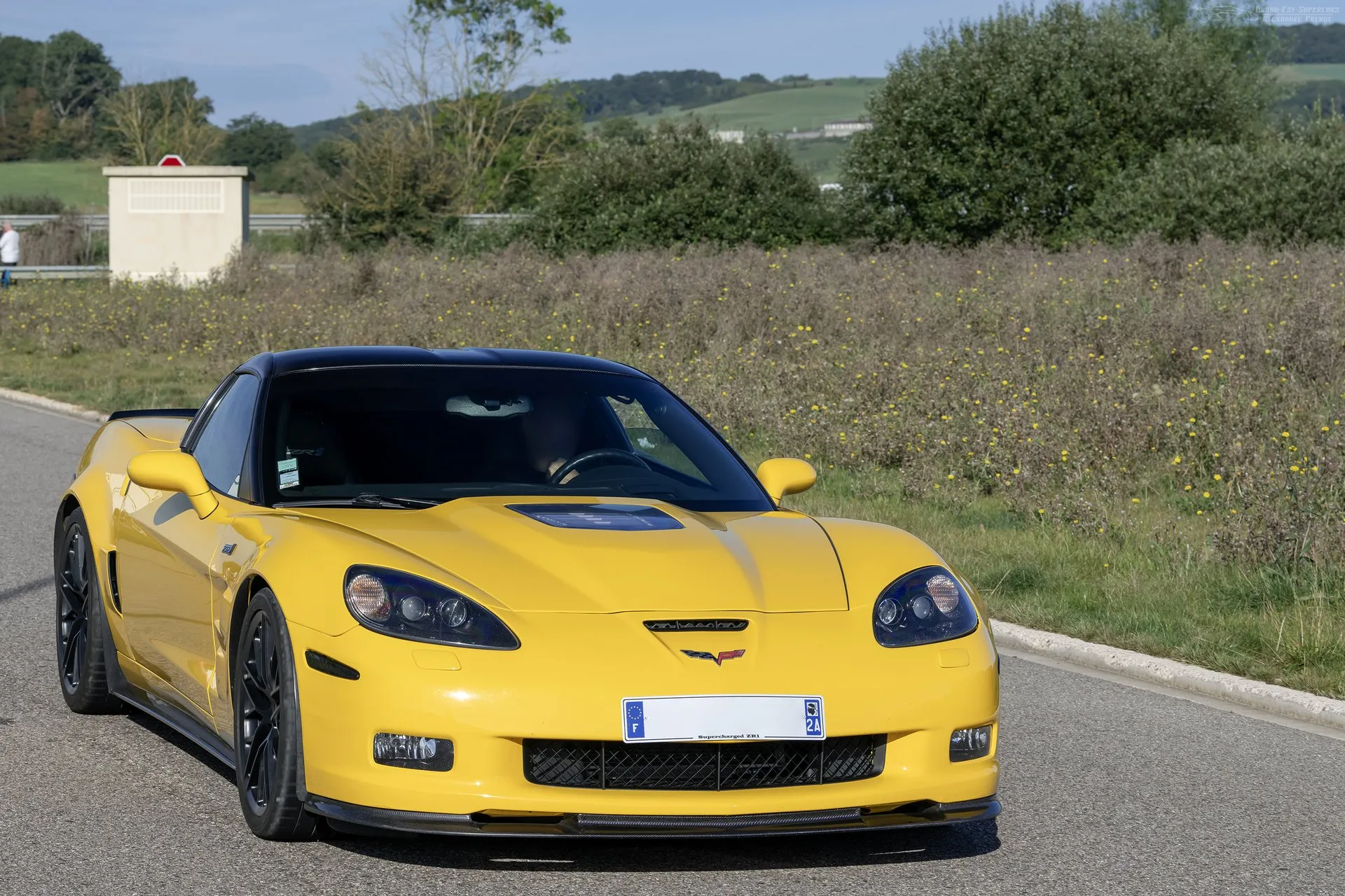 Yellow Corvette ZR1 driving toward the camera on a country road, with a black roof, a clear window in the hood, black multi-spoke wheels and a black splitter under the front bumper.