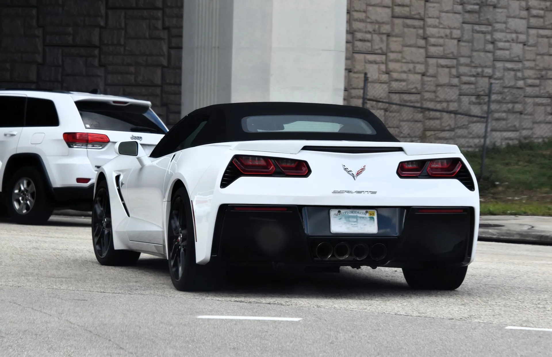 White C7 Corvette Stingray convertible with its black soft top raised and black wheels, on a city street, seen from behind.