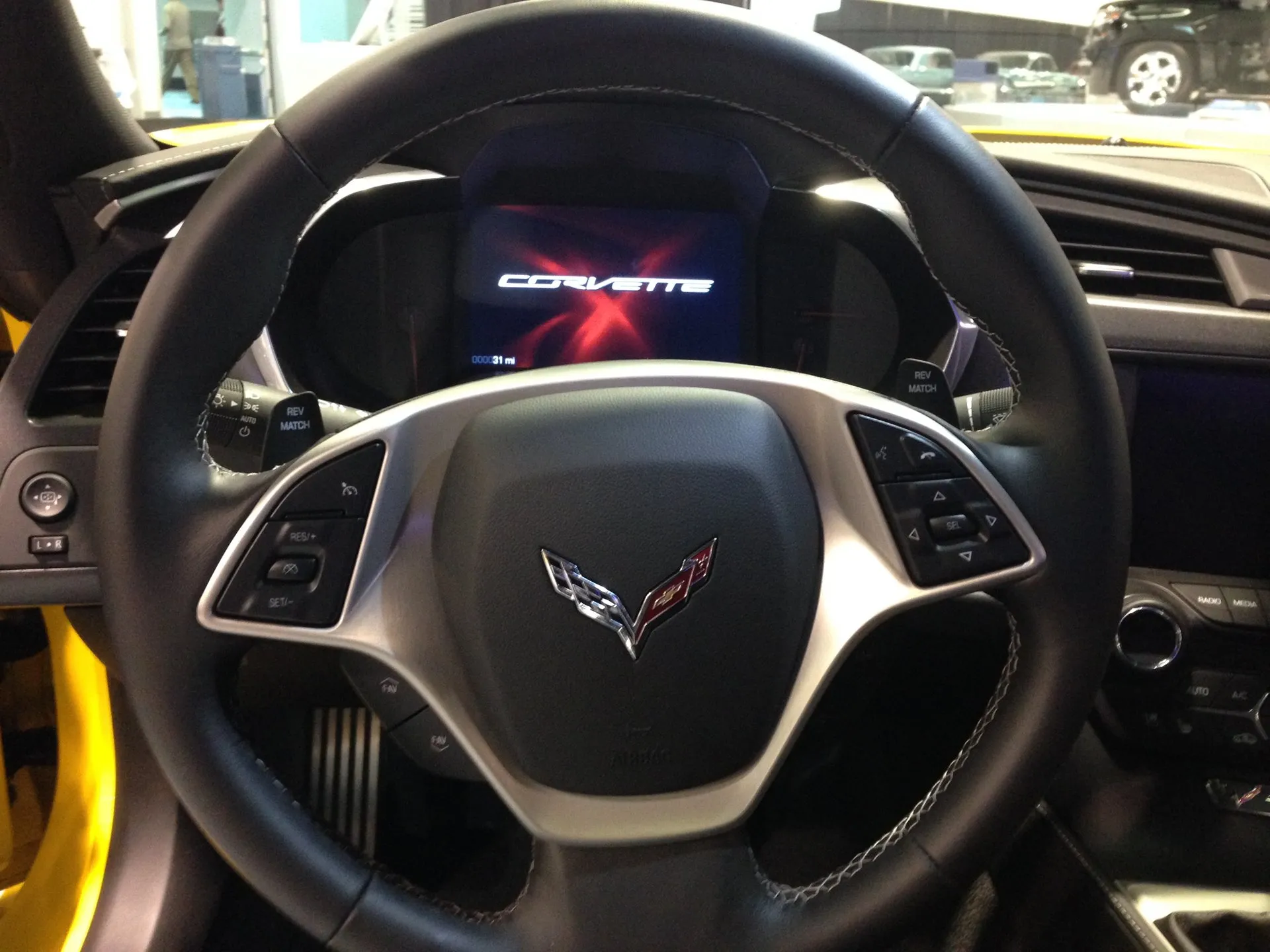 The steering wheel of a yellow C7 Corvette Stingray in a show hall, with paddles marked REV MATCH behind it and the gauge cluster screen showing the Corvette name.