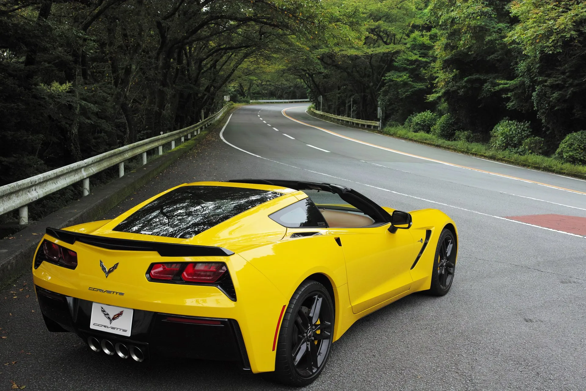 A yellow C7 Corvette Stingray coupe with its roof panel off and a tan interior, seen from behind on the mountain road, showing four center exhaust tips.