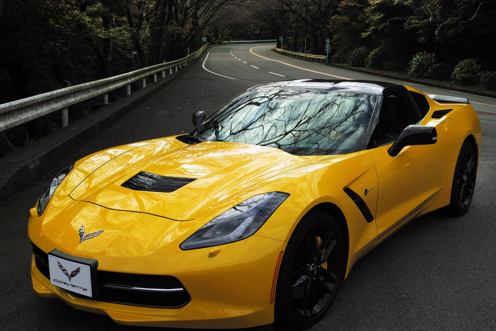 Yellow C7 Corvette Stingray coupe with black wheels and yellow brake calipers parked on a tree-lined mountain road, seen from the front left.