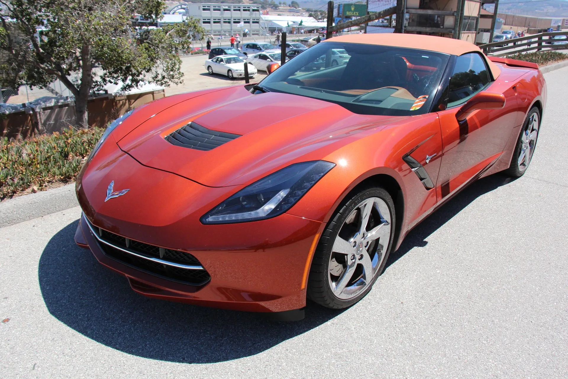 An orange C7 Corvette Stingray convertible with a tan soft top and chrome five-spoke wheels, parked on a road above a racetrack paddock, seen from the front left.