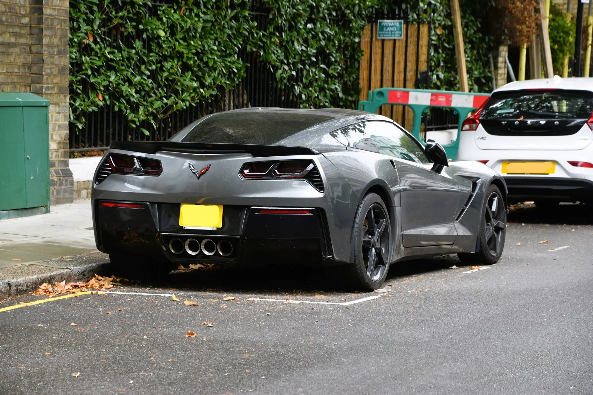 A gray C7 Corvette Stingray coupe with black wheels and a rear spoiler parked at a London curb, seen from behind, showing four center exhaust tips.