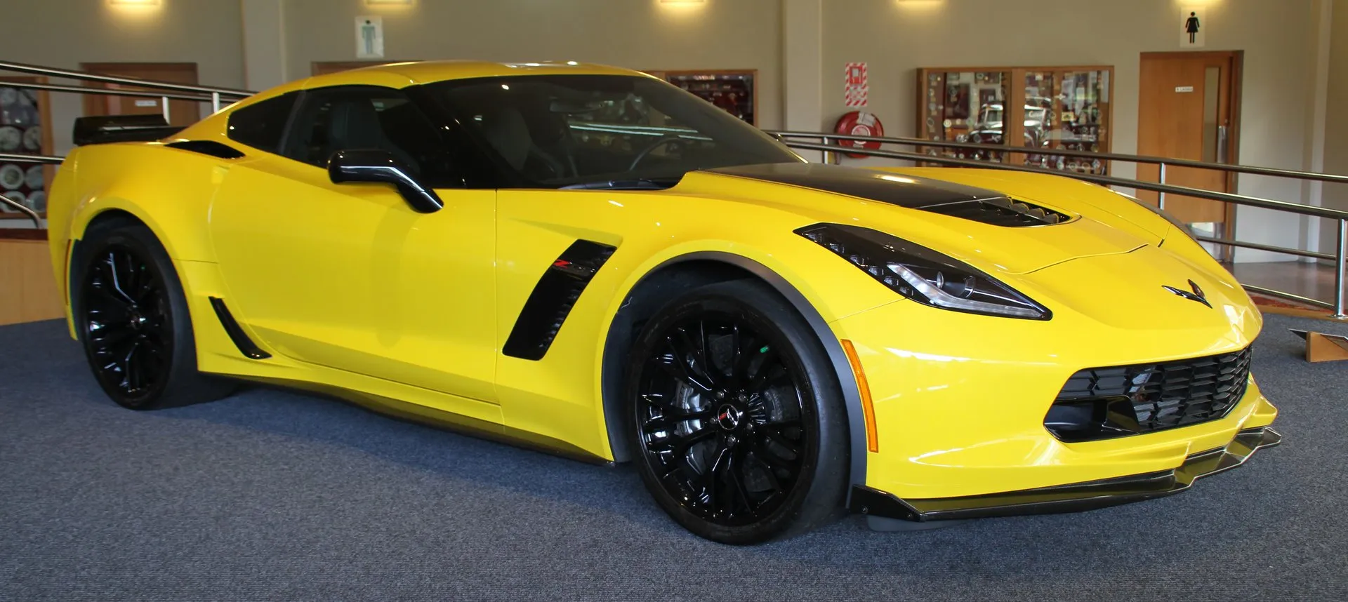 A yellow C7 Corvette Z06 coupe with black wheels, a black hood center and a carbon front splitter, displayed indoors on blue carpet, seen from the front right.