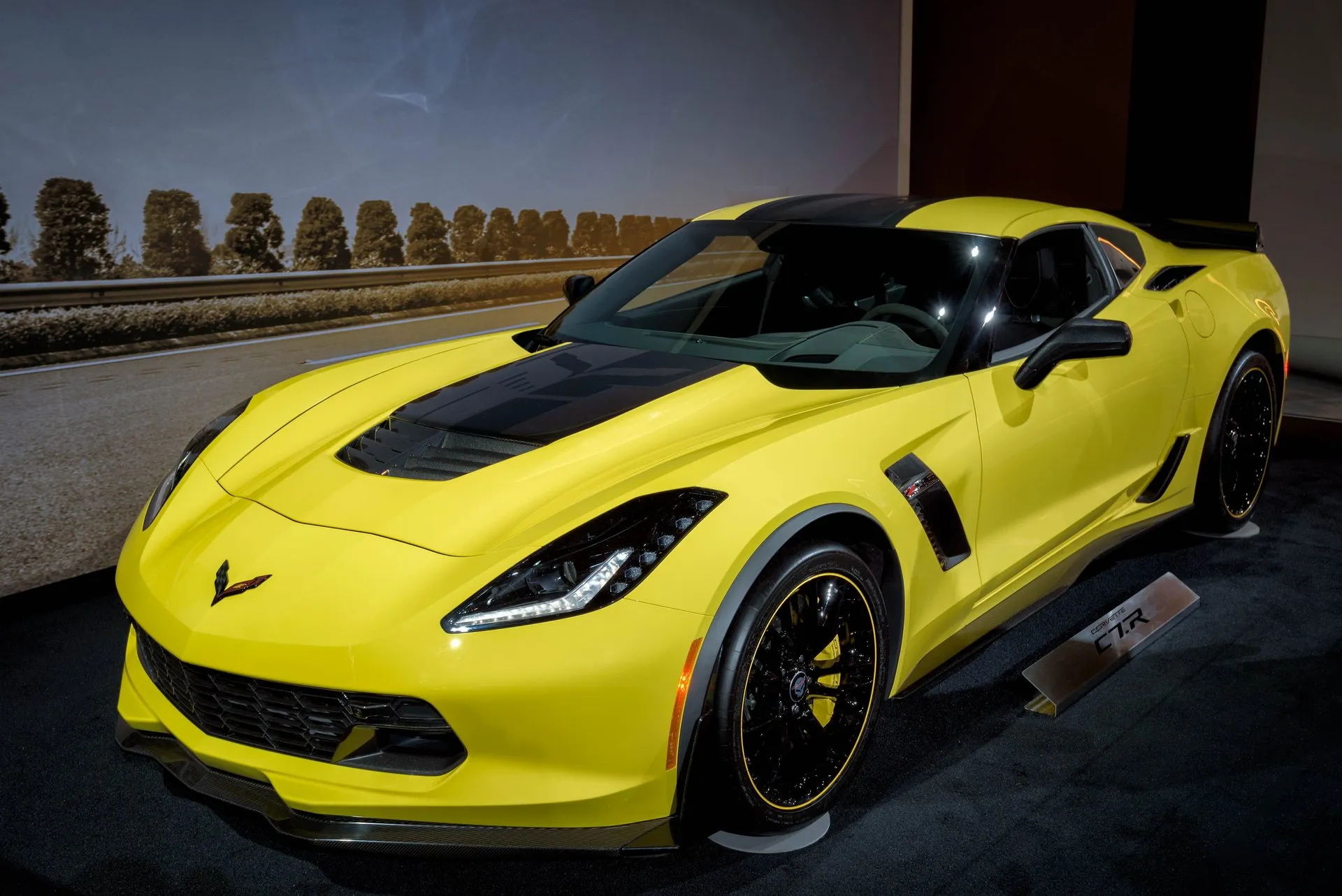 Yellow Corvette Z06 C7.R Edition coupe with a black hood center, black wheels with a thin yellow rim stripe and yellow brake calipers, on a display stand at an auto show beside a C7.R placard, seen from the front left.