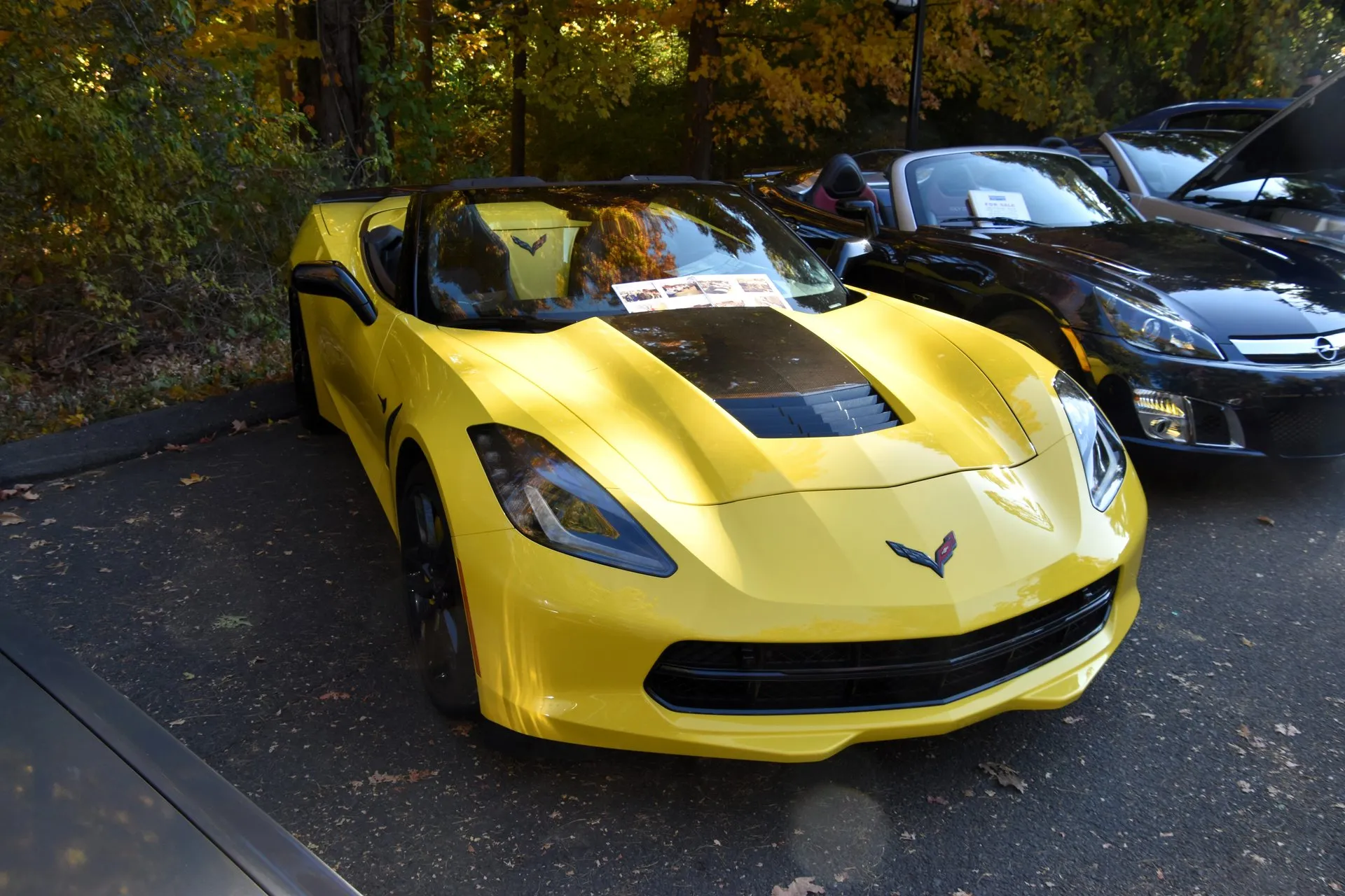Yellow C7 Corvette Stingray convertible with its top down, a black hood vent and black wheels, at an outdoor car show under autumn trees, seen from the front.