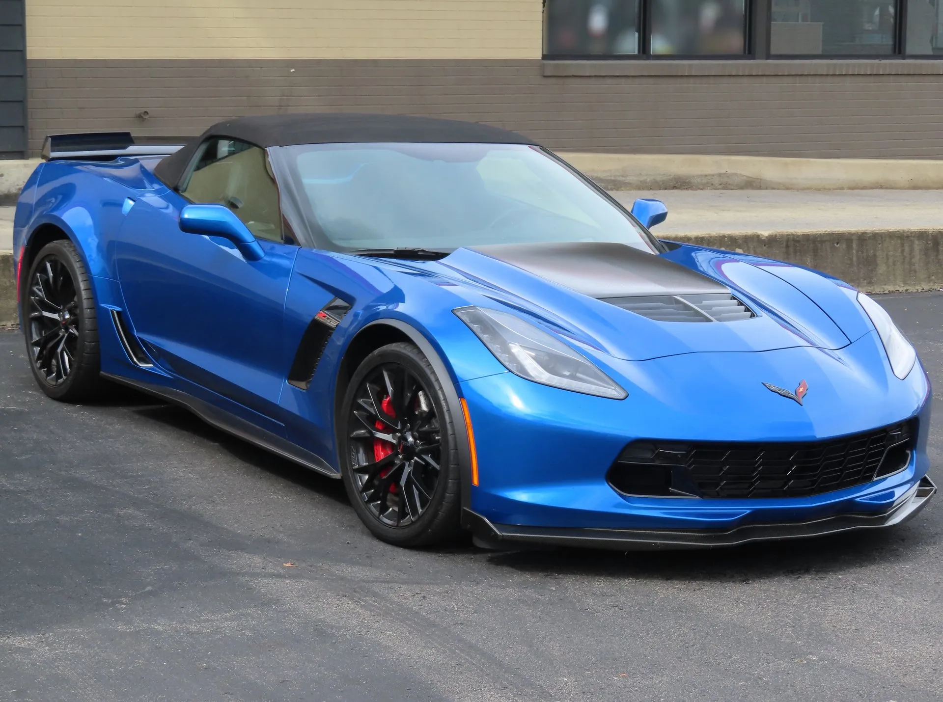 Blue 2016 Chevrolet Corvette Z06 convertible with its black soft top raised, a dark vented hood panel, black wheels and red brake calipers, parked on asphalt, seen from the front right.