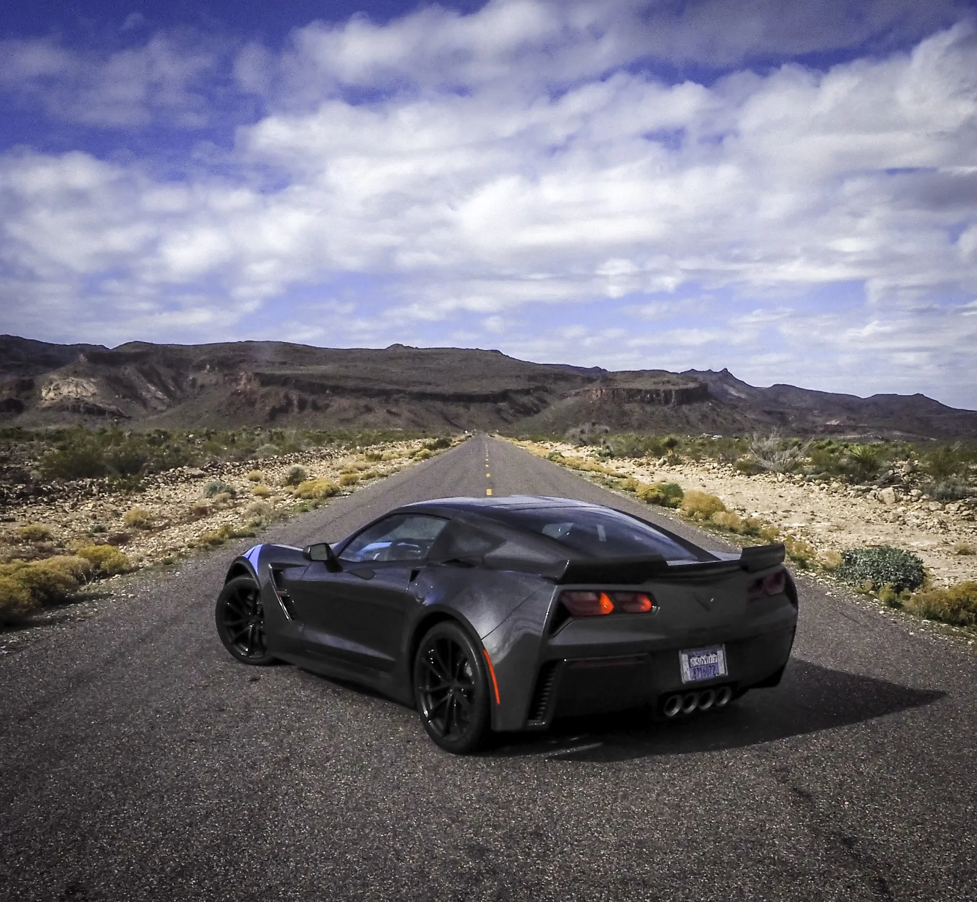 The dark gray Grand Sport from behind on an empty desert highway, showing its wide rear fenders, rear spoiler and four center exhaust tips.