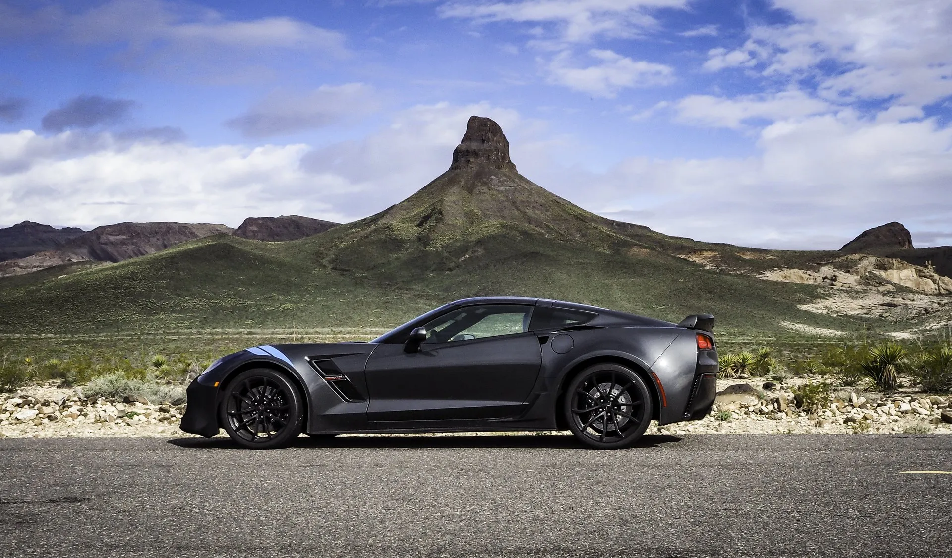 Dark gray Corvette Grand Sport coupe with black wheels, light blue hash marks on the front fender and a black cover over its nose, in profile on a desert road below a rocky peak.