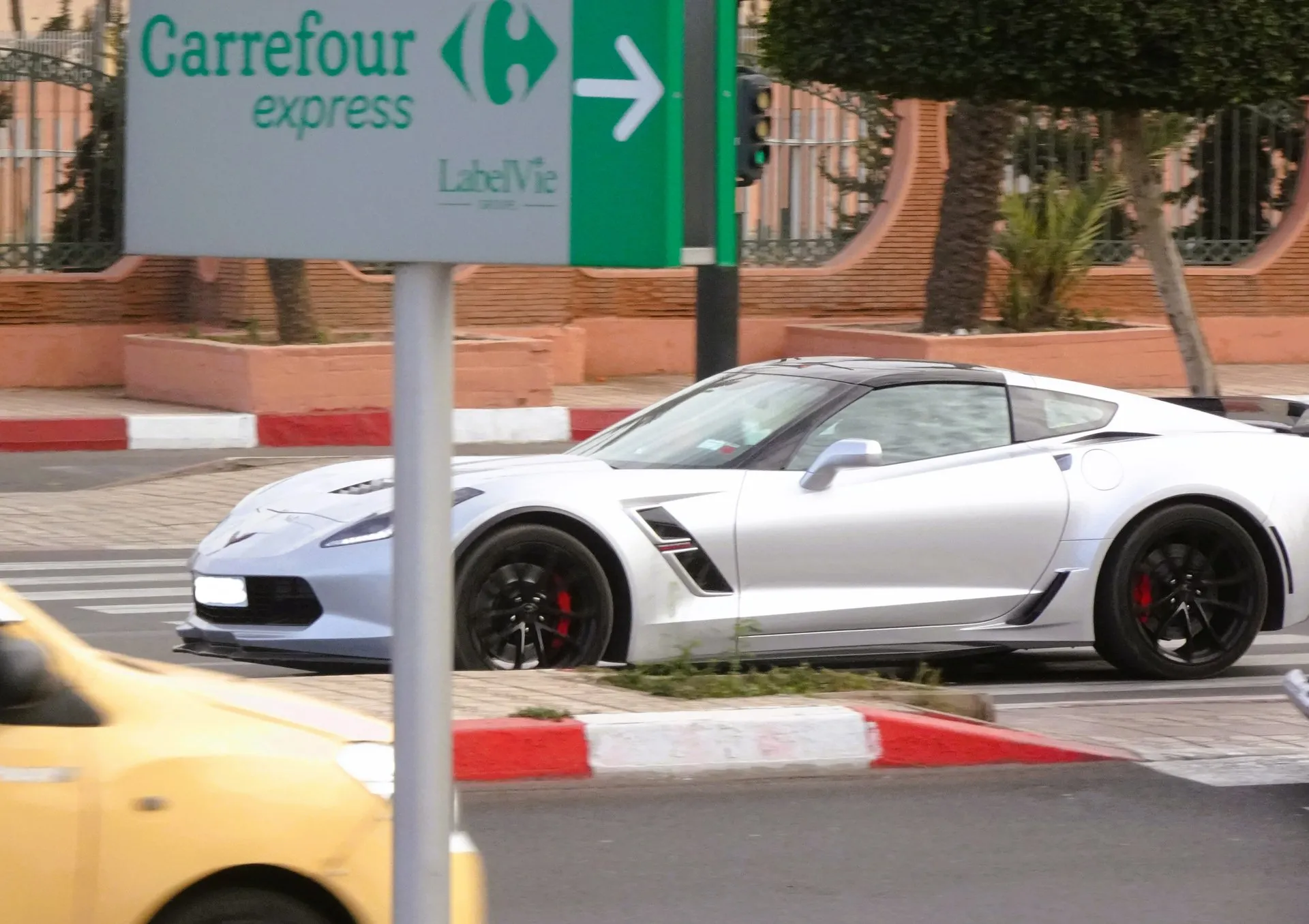Silver C7 Corvette Grand Sport coupe with a black roof, black wheels and red brake calipers on a city street, behind a store sign, with a yellow taxi in the foreground.