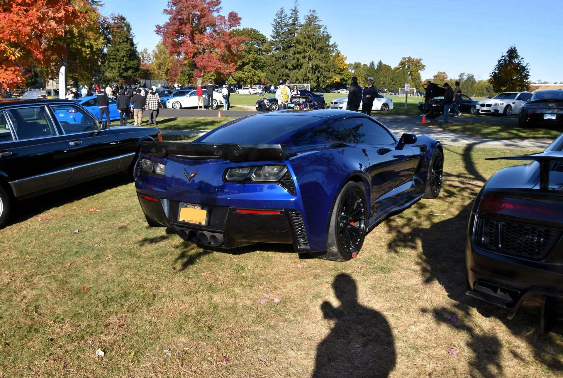 Blue C7 Corvette Z06 coupe with black wheels and a rear spoiler, seen from the rear right on the grass at a fall car show, with other cars and people behind it.