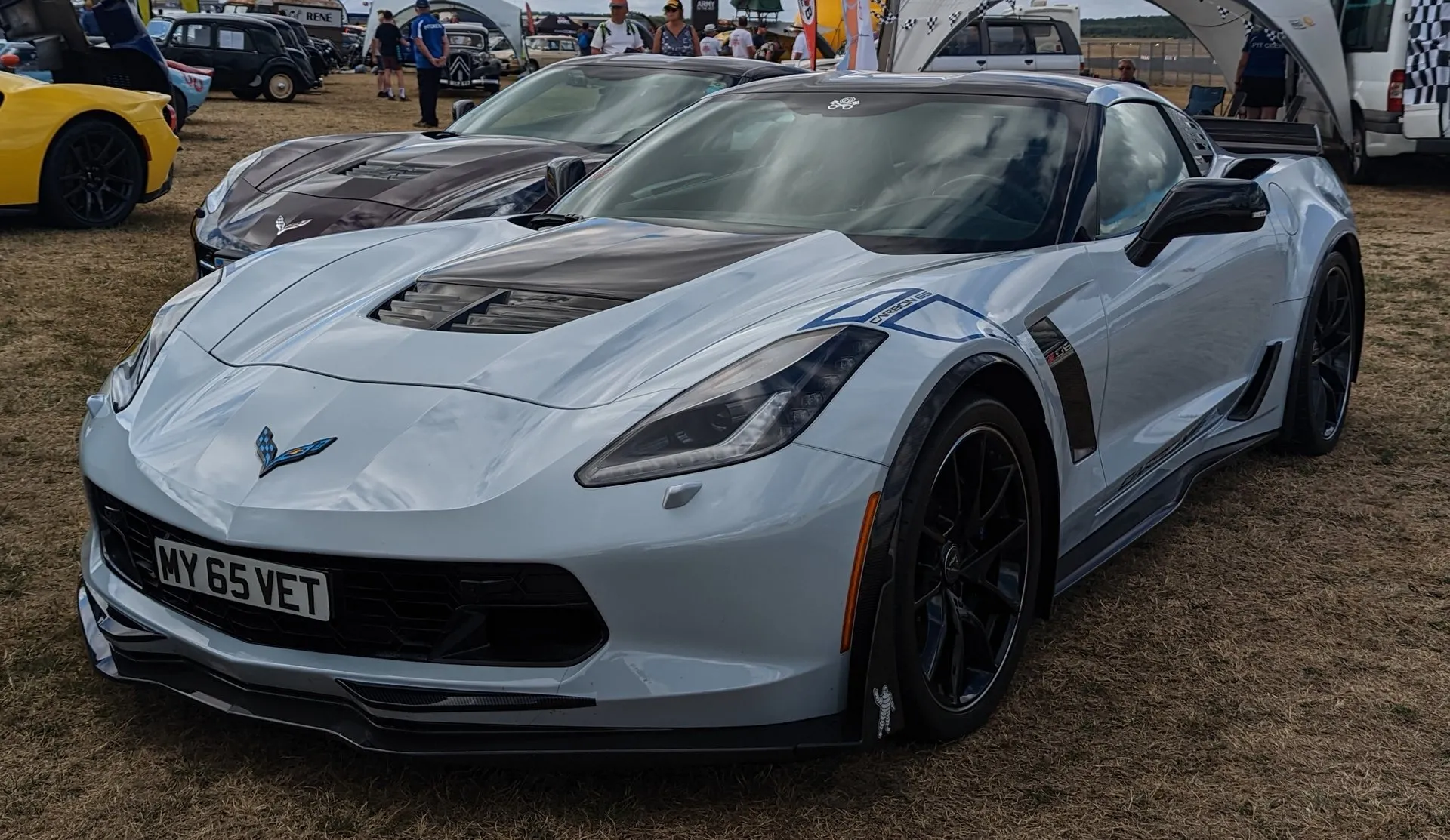 A light gray C7 Corvette coupe with a visible carbon hood section, blue graphics on the front fender and black wheels, parked on grass at a car show beside a darker Corvette.