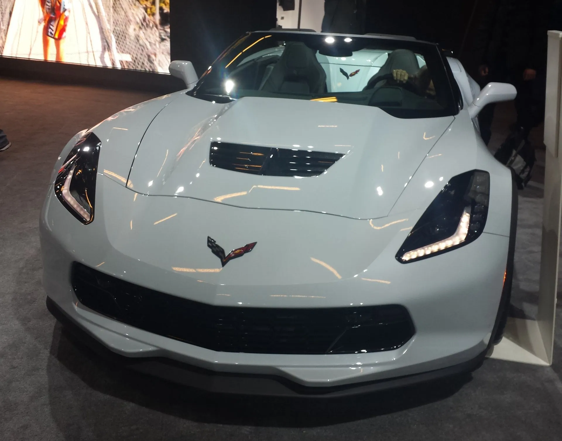 A pale gray C7 Corvette convertible with a wide front bumper and a small hood vent, seen head-on on an auto show stand.