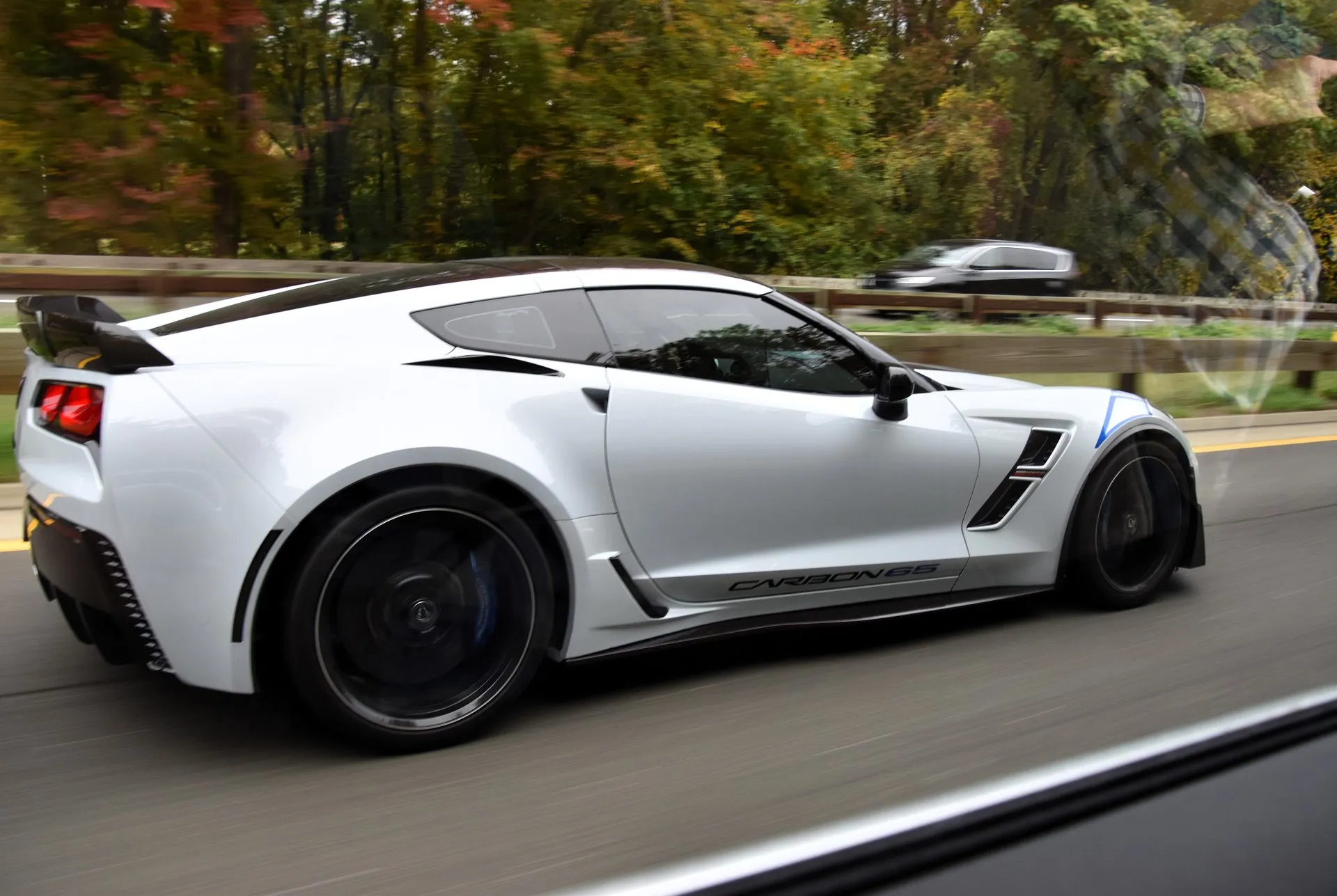 A very light gray C7 Corvette coupe on a tree-lined highway, photographed from a moving car: Carbon 65 script low on the door, a blue stripe on the front fender, a carbon rear spoiler and black wheels.