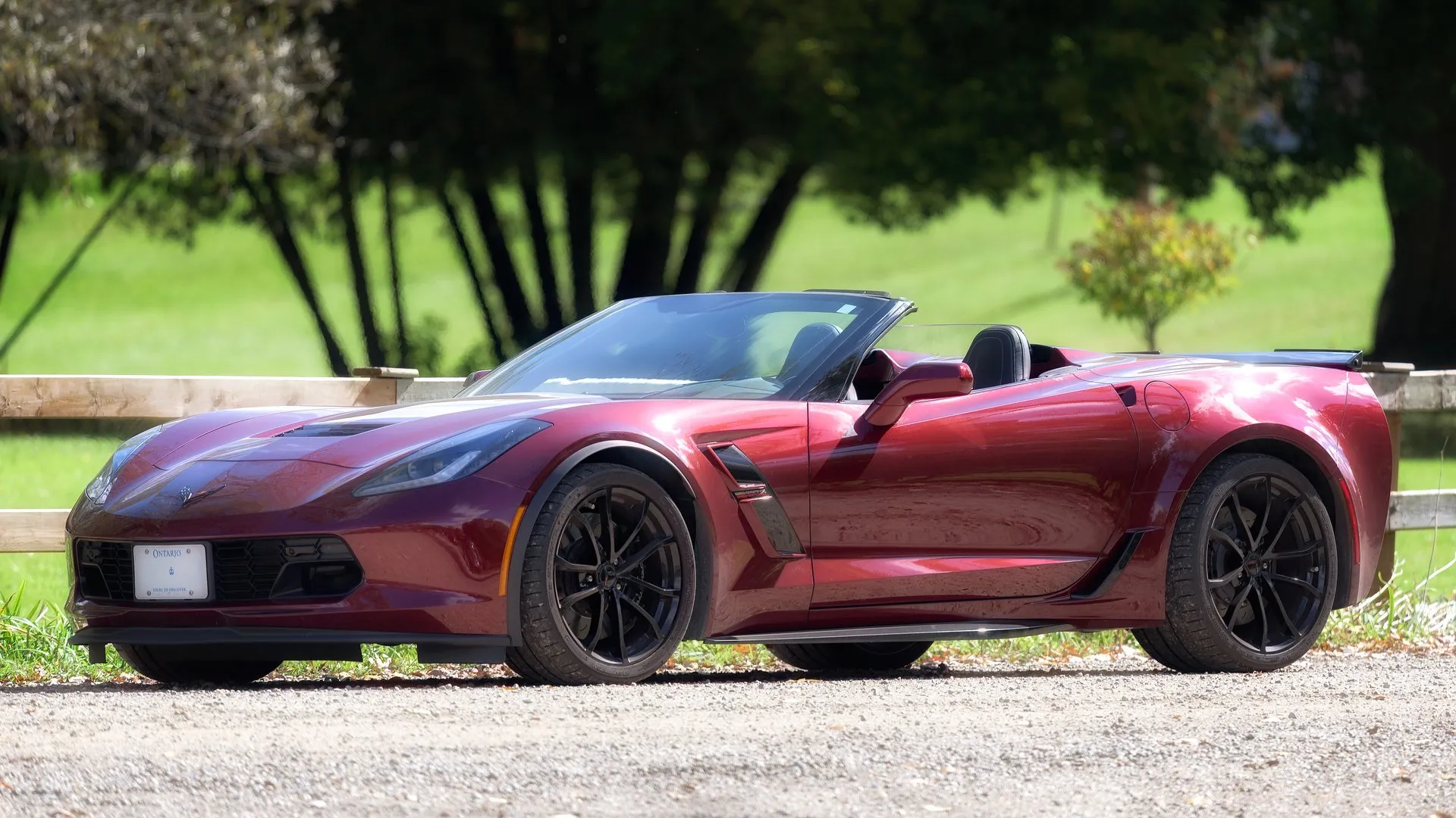 Dark red Chevrolet Corvette Grand Sport convertible with its top down and black wheels, parked on gravel beside a wooden fence, seen from the front left.