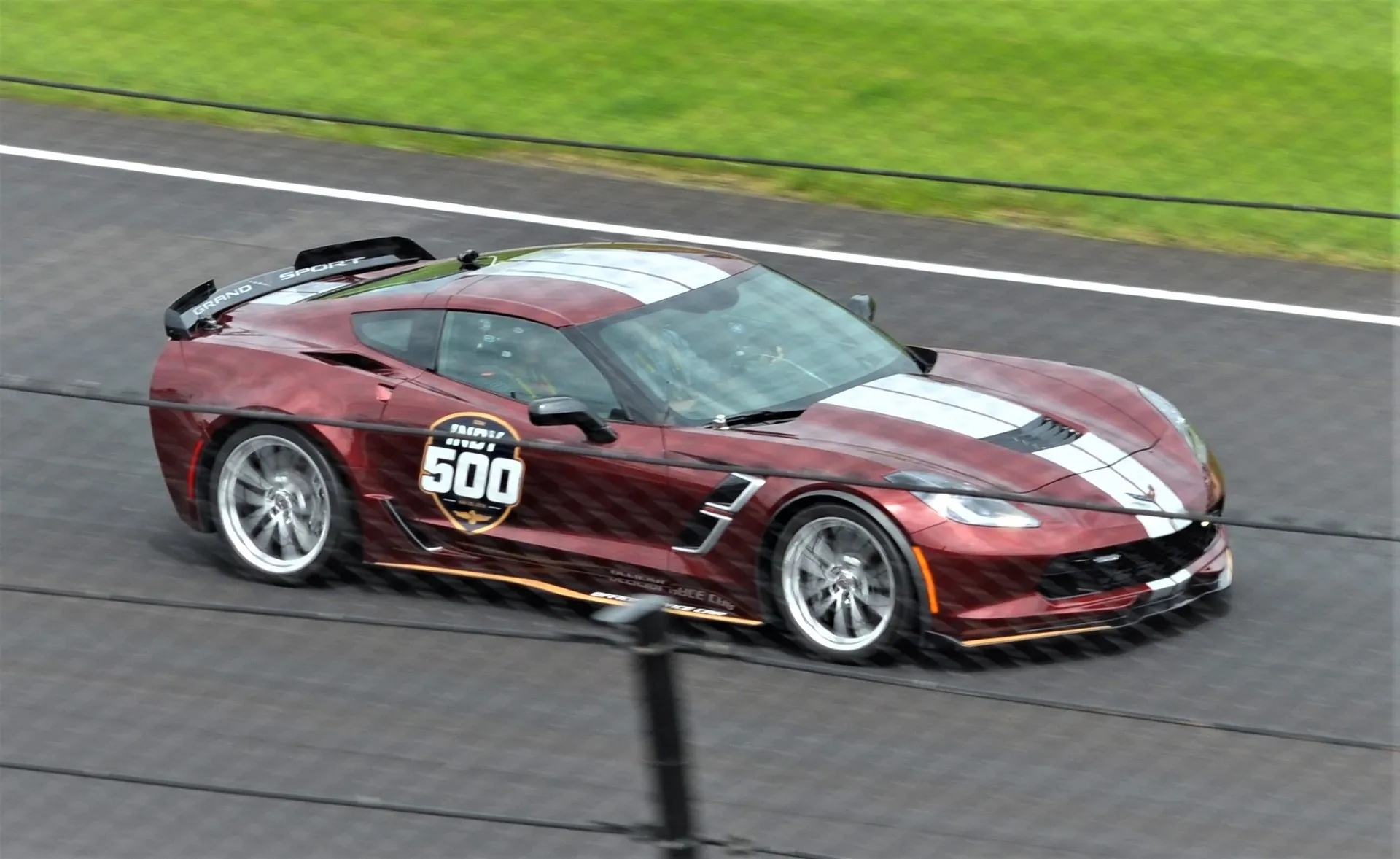 Dark red Chevrolet Corvette Grand Sport coupe with wide silver stripes over the hood and roof and an Indy 500 logo on the door, driving on the Indianapolis track, seen through a catch fence.