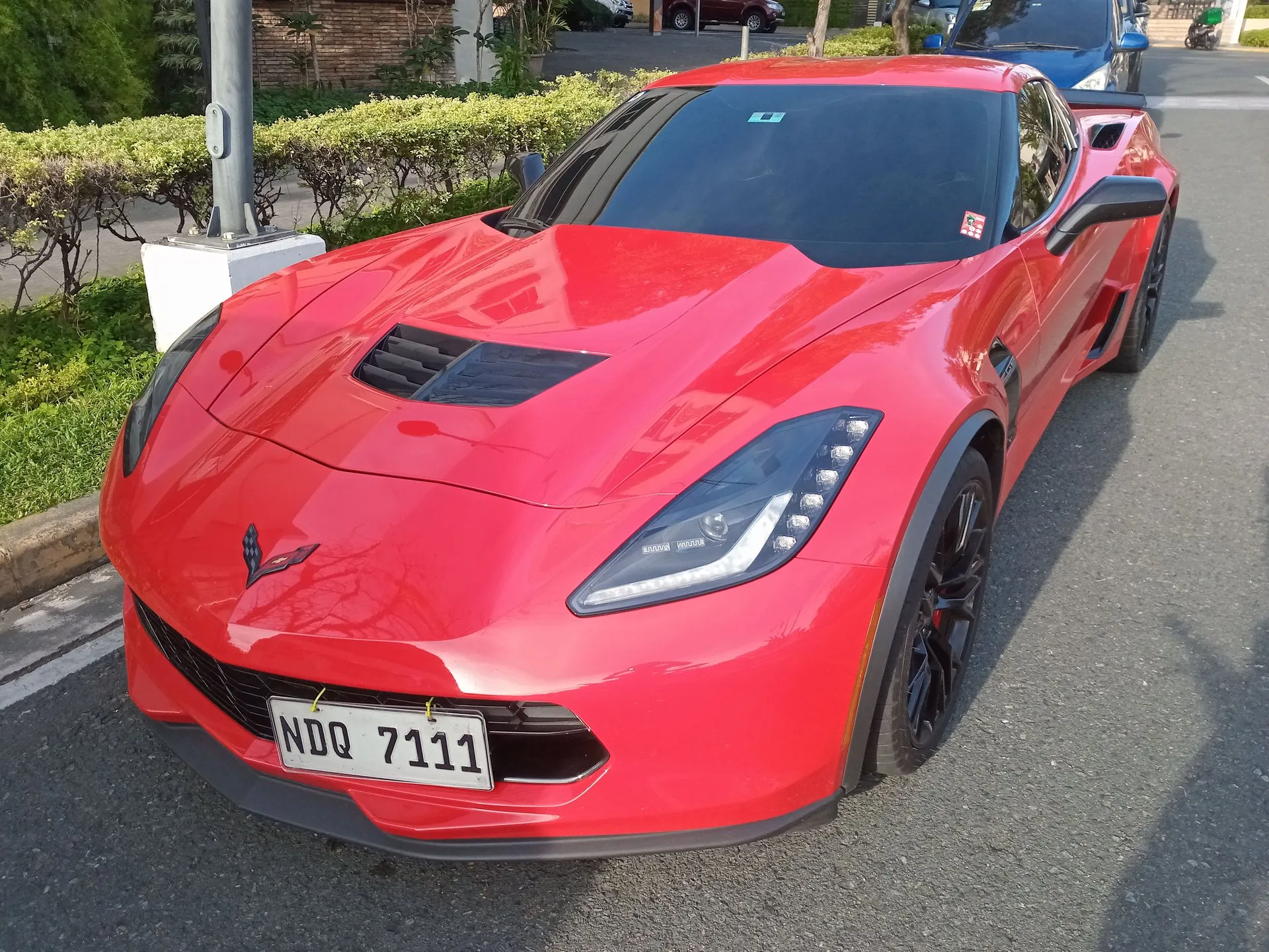 Red C7 Chevrolet Corvette Z06 coupe with a vented hood, black wheels and red brake calipers, parked at a curb, seen from the front left.