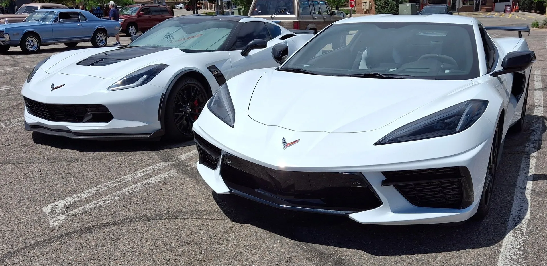 A white C7 Corvette Z06 with a black hood vent, black wheels and red brake calipers parked beside a white C8 Corvette Stingray with a tall rear wing, both seen from the front in a car show parking lot.
