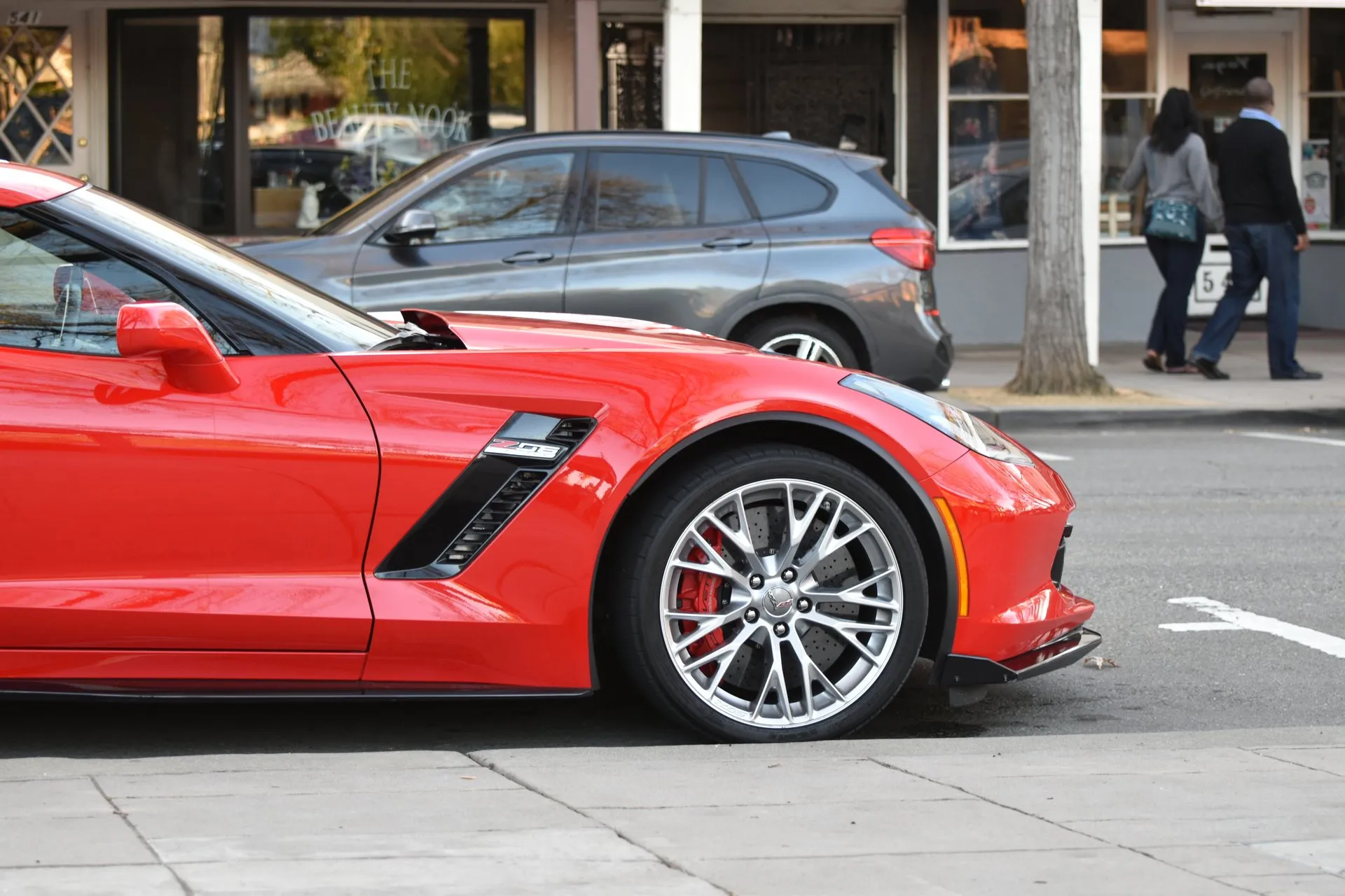 Front wheel of a red C7 Corvette Z06 parked at a curb: a silver multi-spoke wheel over a red brake caliper and a cross-drilled rotor, below the Z06 badge in the front fender vent.