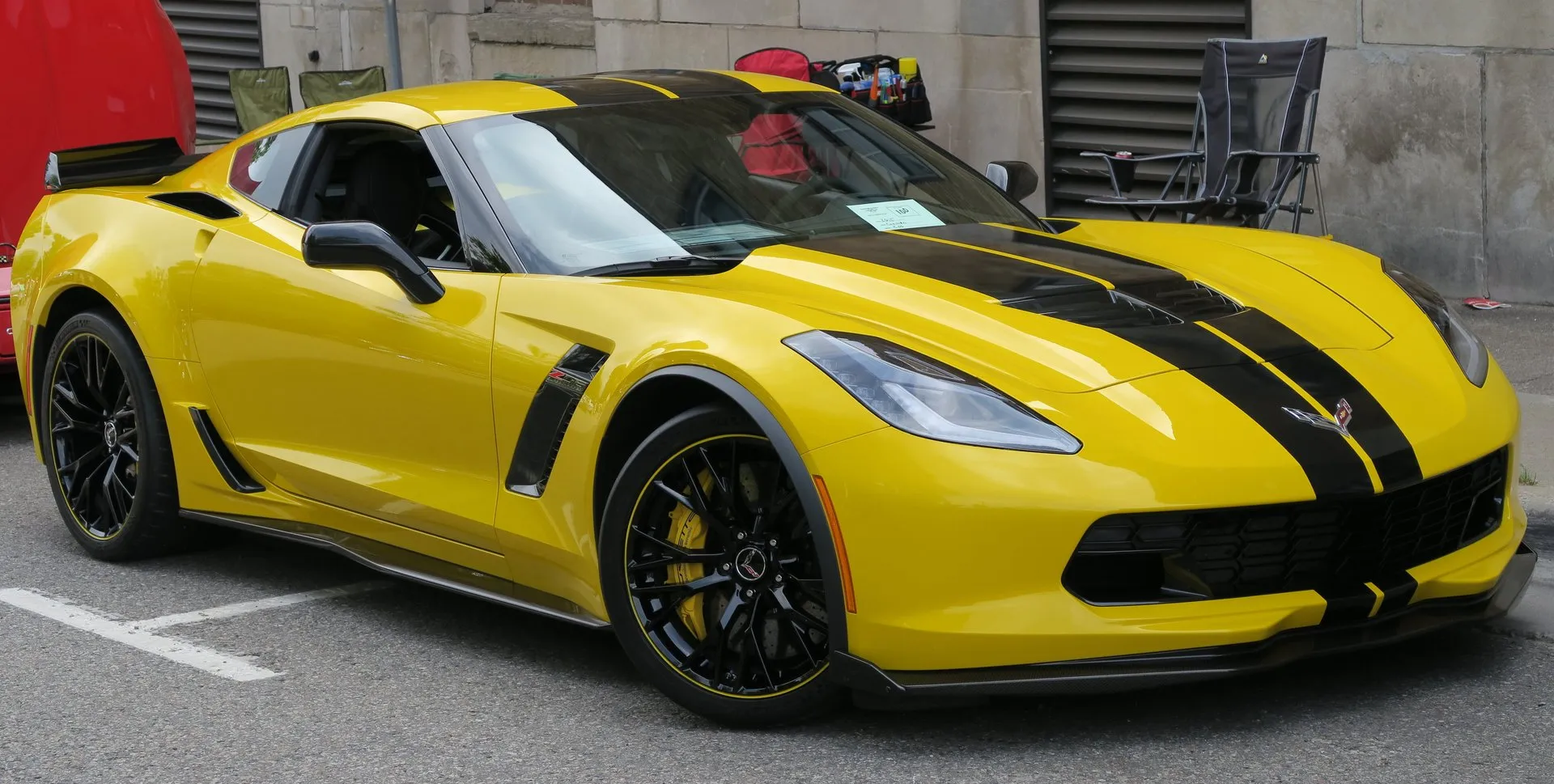 Yellow C7 Chevrolet Corvette Z06 coupe with black stripes over the hood and roof, black wheels with a yellow rim stripe and yellow brake calipers, parked at a street car show, seen from the front right.