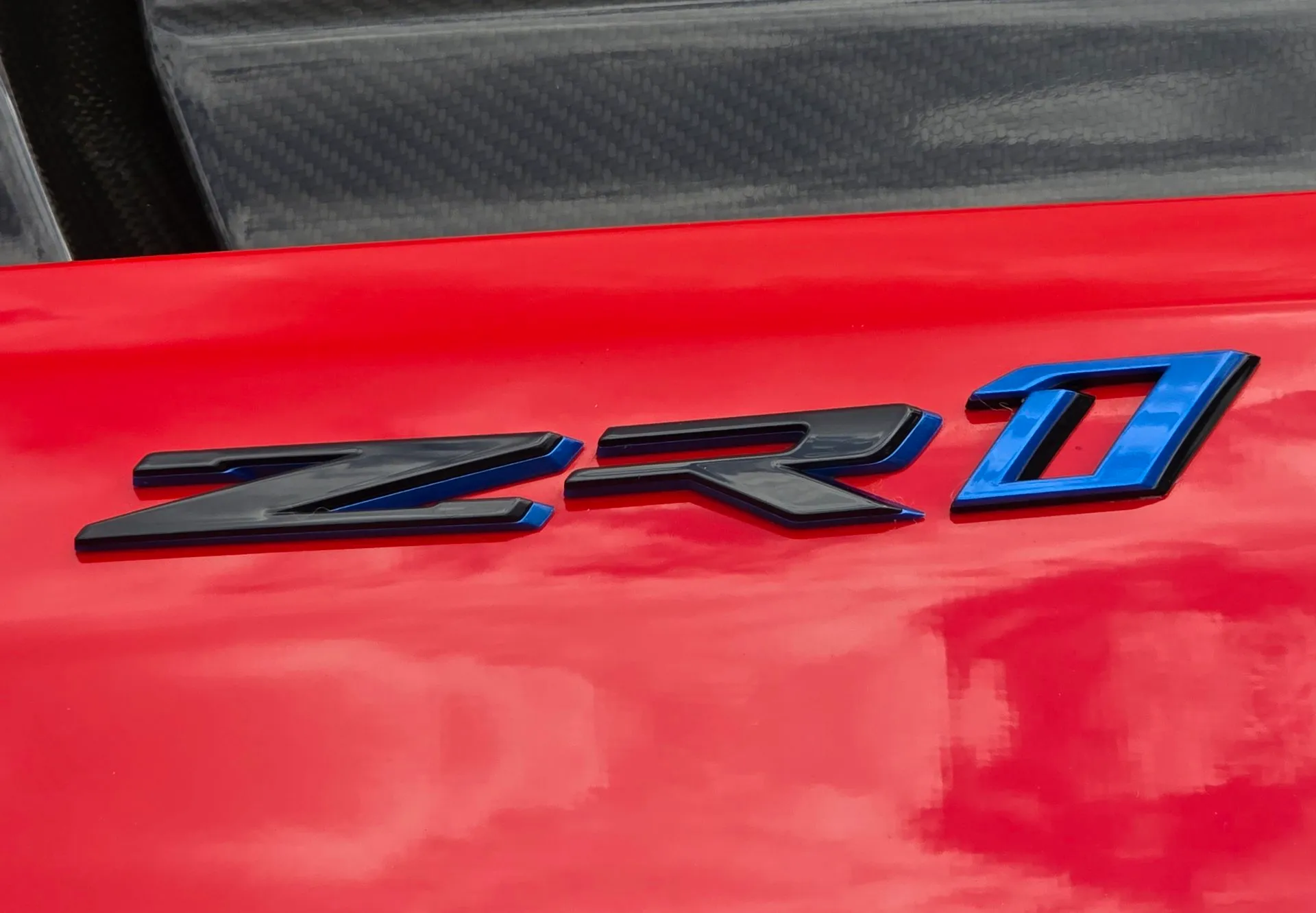 Close-up of a ZR1 badge on a red Corvette: a black Z and R and a blue 1, below a panel of bare carbon fiber.