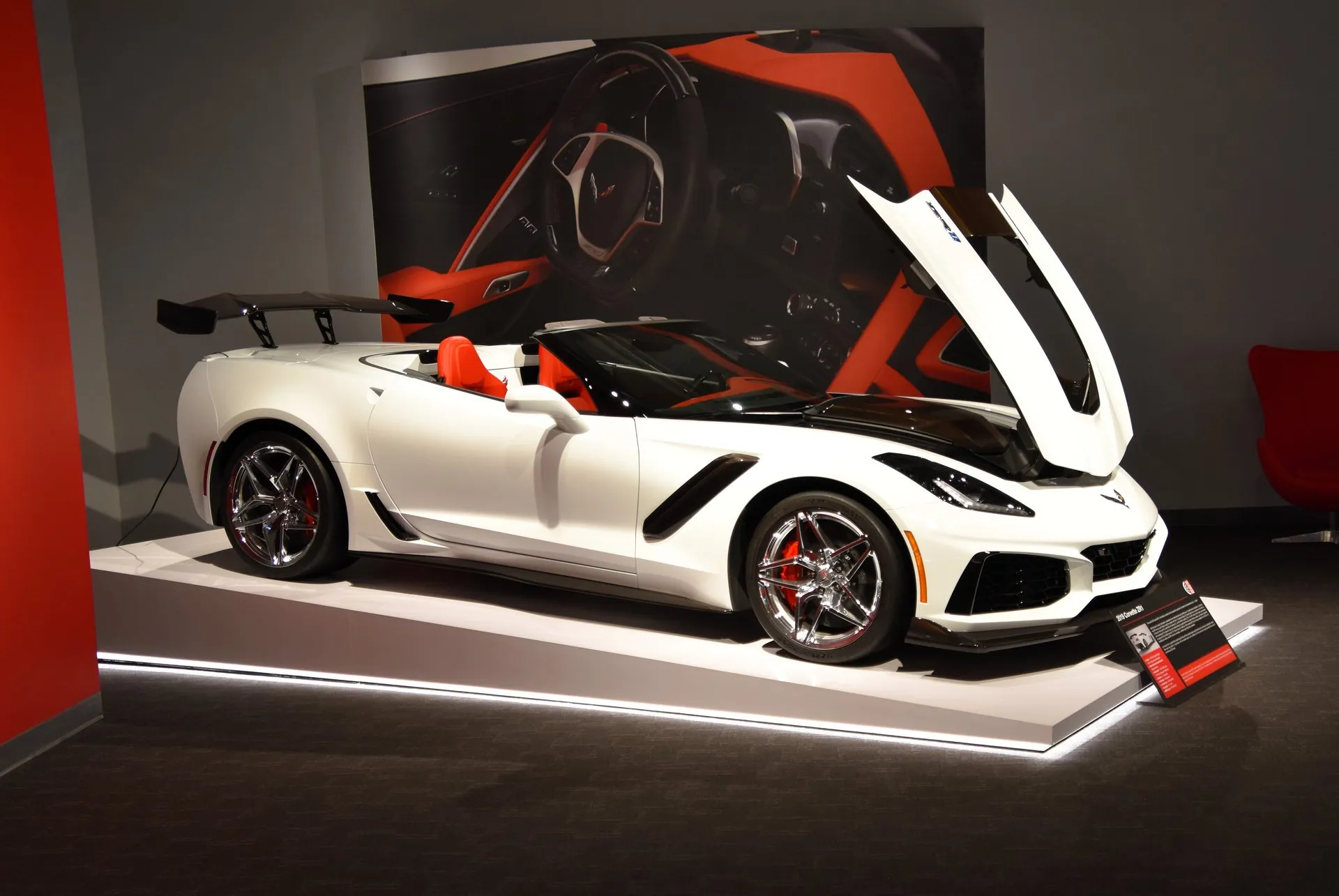 A white Corvette ZR1 convertible on a museum display platform, seen from the front right with its hood raised and top down, showing a tall rear wing, chrome wheels, red brake calipers and red seats.