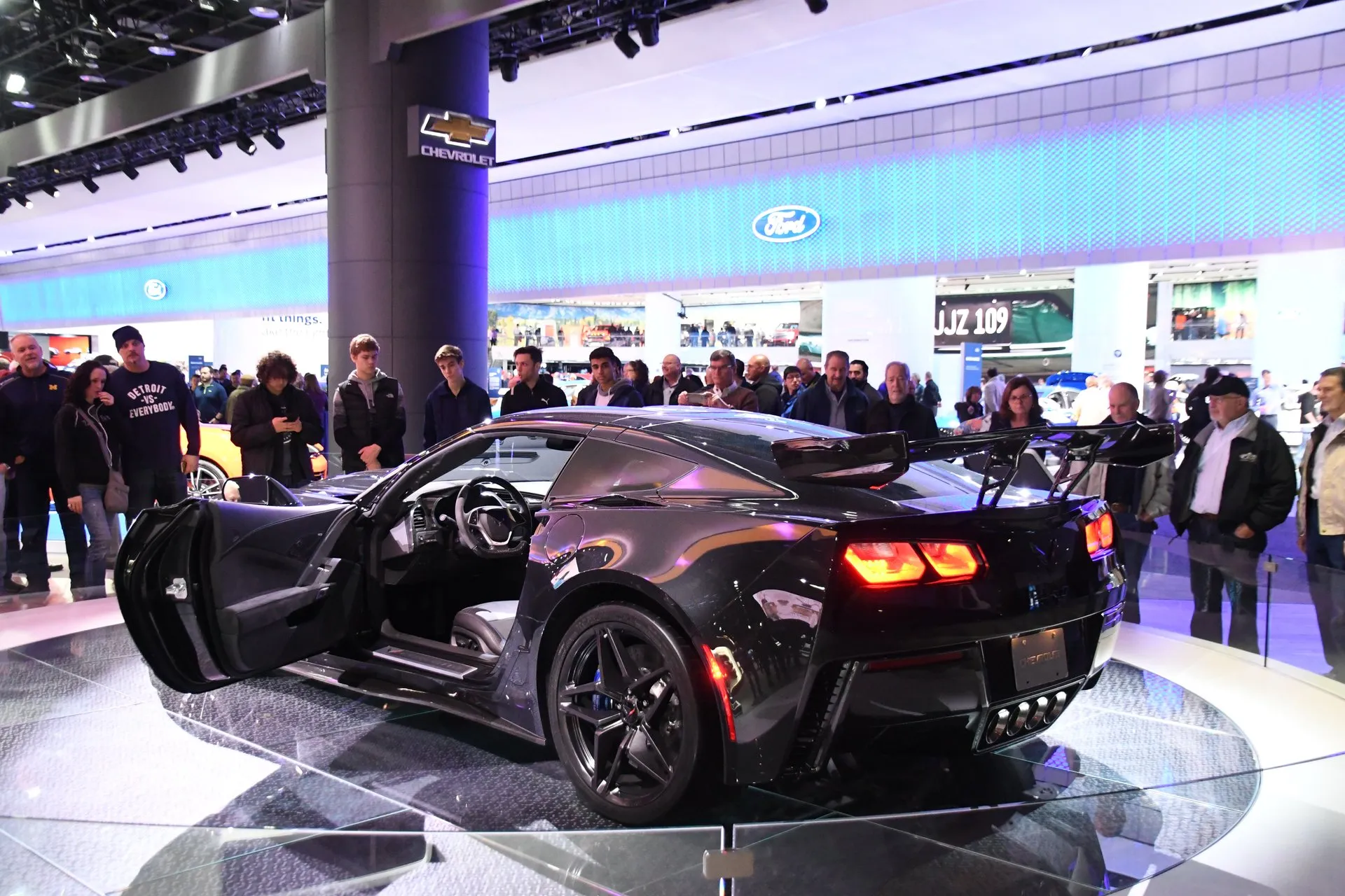 A dark Corvette ZR1 coupe on a round display stand at an auto show, seen from the rear left with its door open, showing a tall rear wing on stanchions and four exhaust tips in the center, with visitors standing around it.