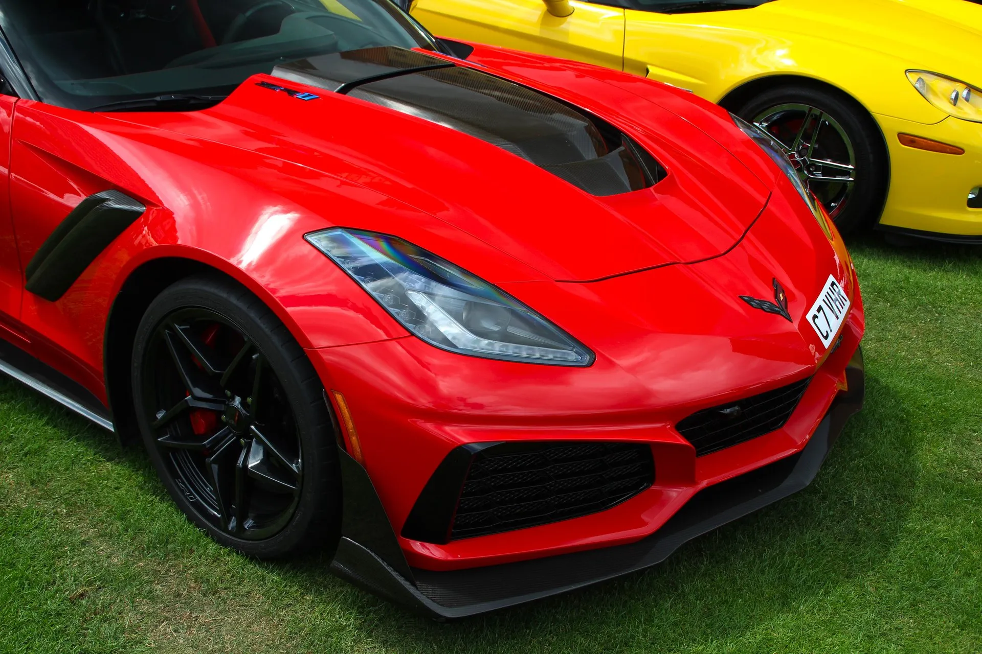 Close view of the front of a red Corvette ZR1 parked on grass, showing the black carbon center of its hood, large grilles in the front fascia, a carbon splitter under the bumper, a black fender vent and black wheels with red brake calipers.