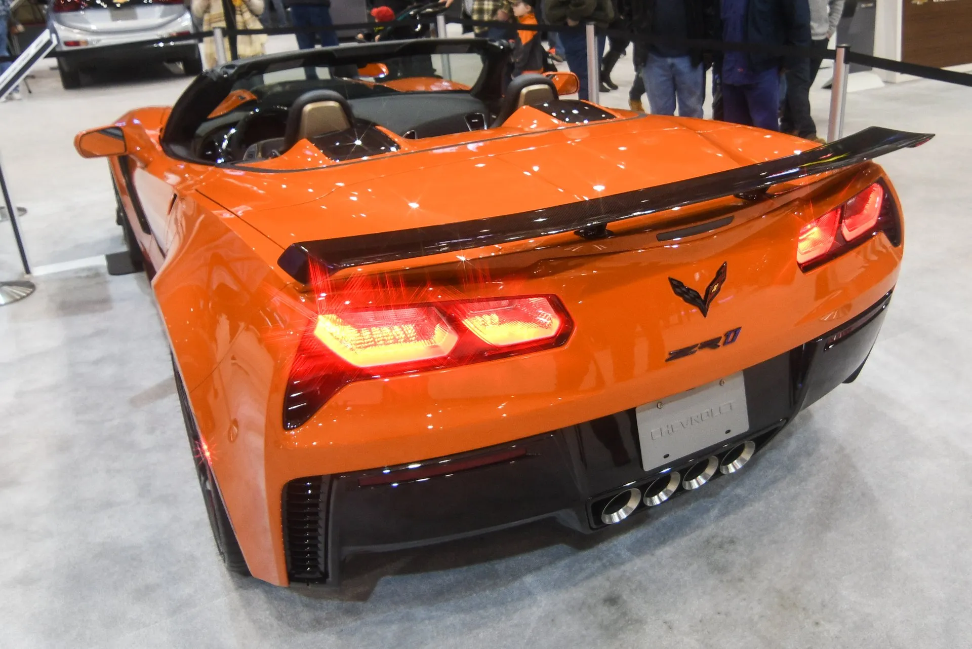 An orange Corvette ZR1 convertible at an auto show, seen from the rear left with its top down, a ZR1 badge on the rear panel, four exhaust tips in the center and a low black wing on short stanchions across the rear deck.