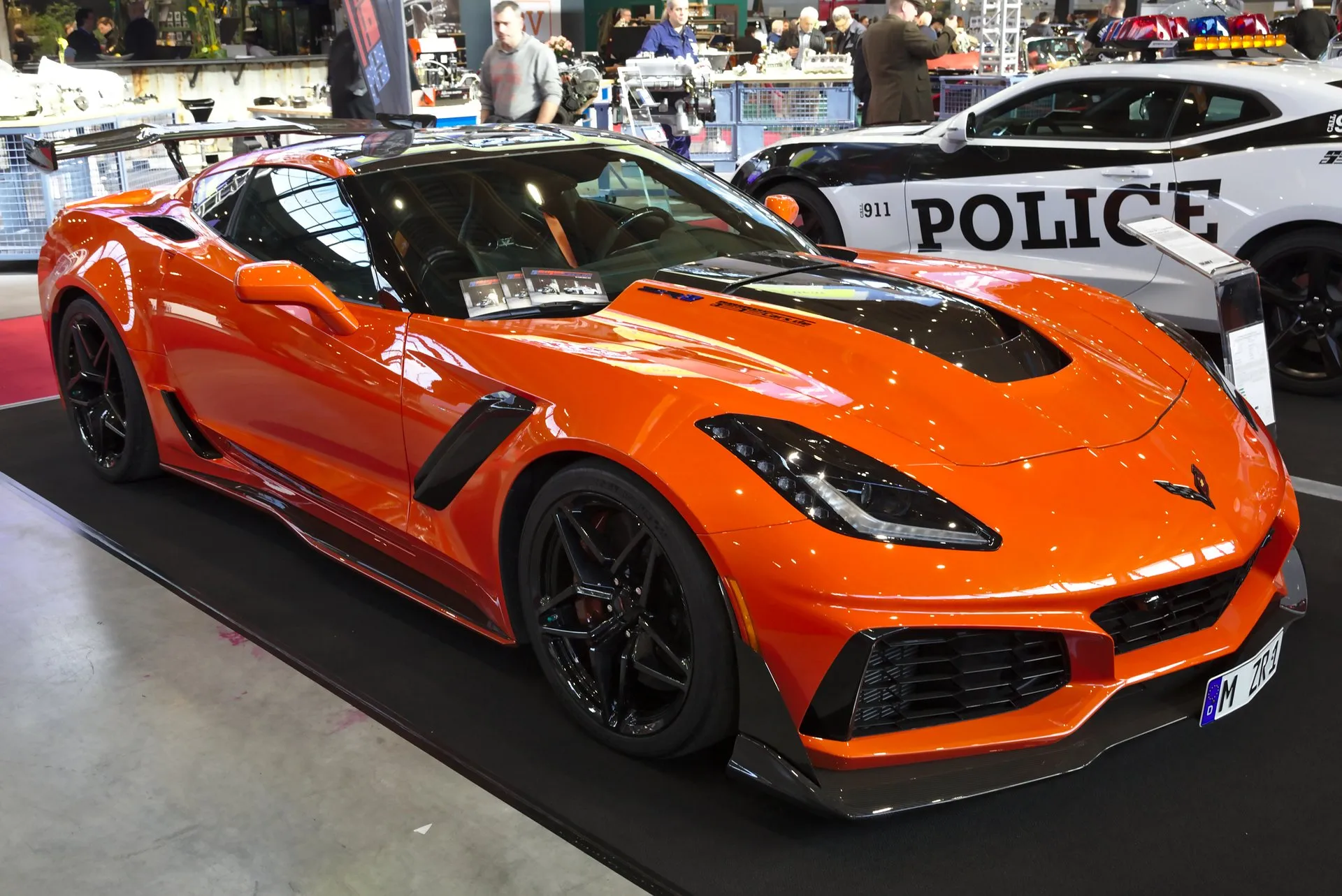 An orange 2019 Chevrolet Corvette ZR1 coupe on a show stand, seen from the front right, with a black center section in its hood, a carbon front splitter, black wheels and a tall rear wing on stanchions.