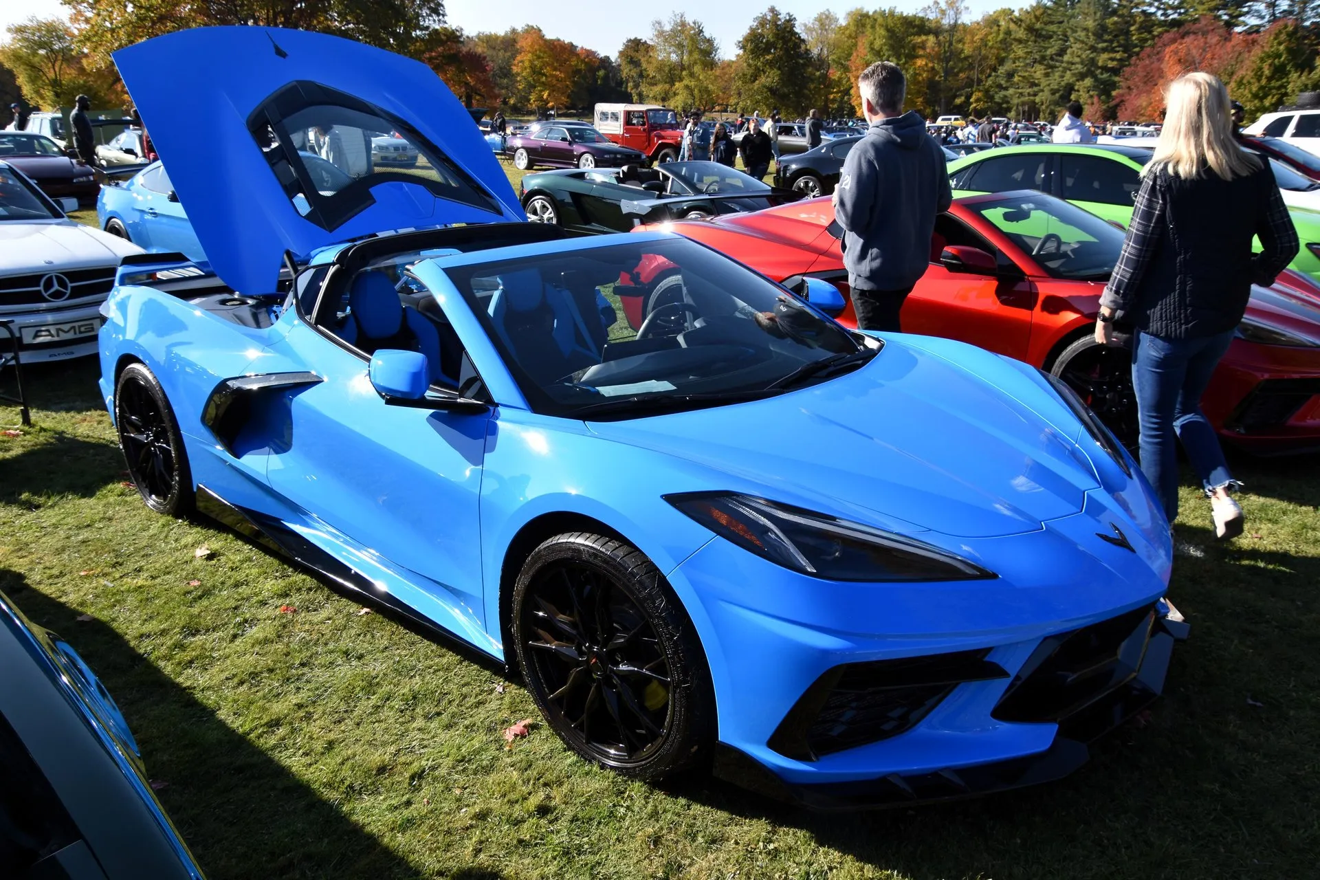 A blue 2023 C8 Corvette Stingray coupe on grass at a car show with its roof panel removed and its glass rear hatch raised behind the cabin, seen from the front right, with spectators walking past.