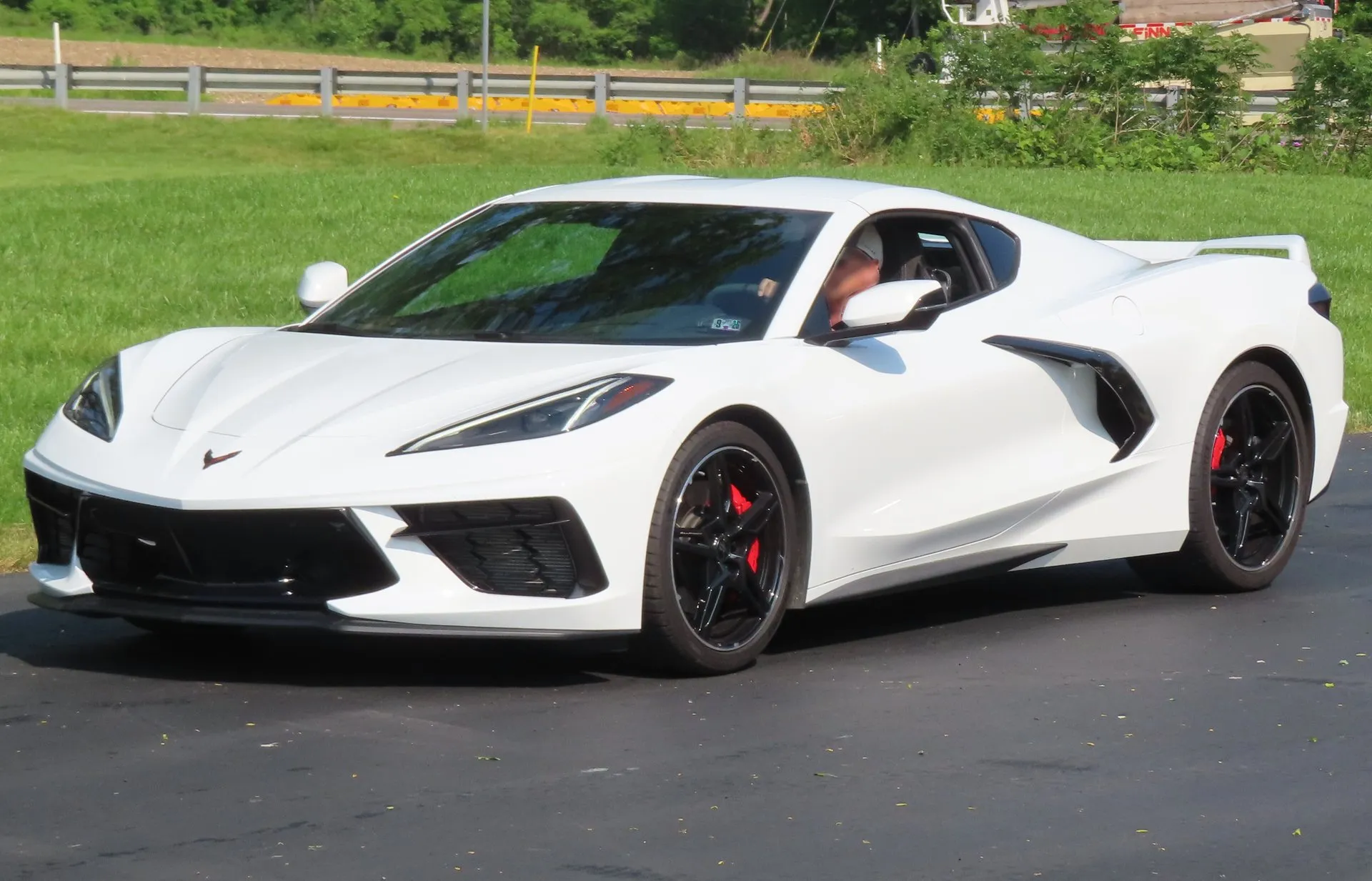 A white 2023 C8 Corvette Stingray coupe with black wheels and red brake calipers on a paved lot beside grass and a guardrail, seen from the front left.